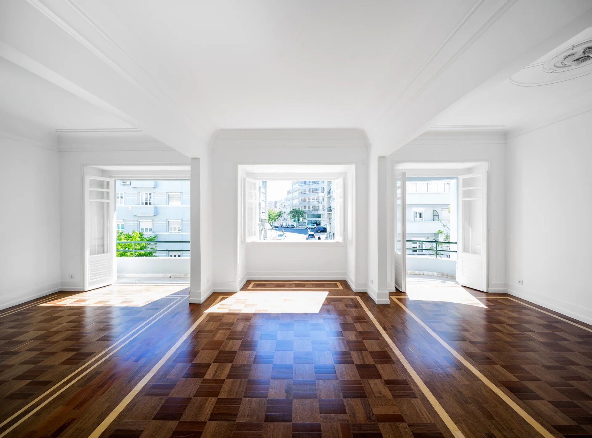 An empty white room with dark parquet hardwood floors featuring light wood borders, and three large windows letting in bright natural light.