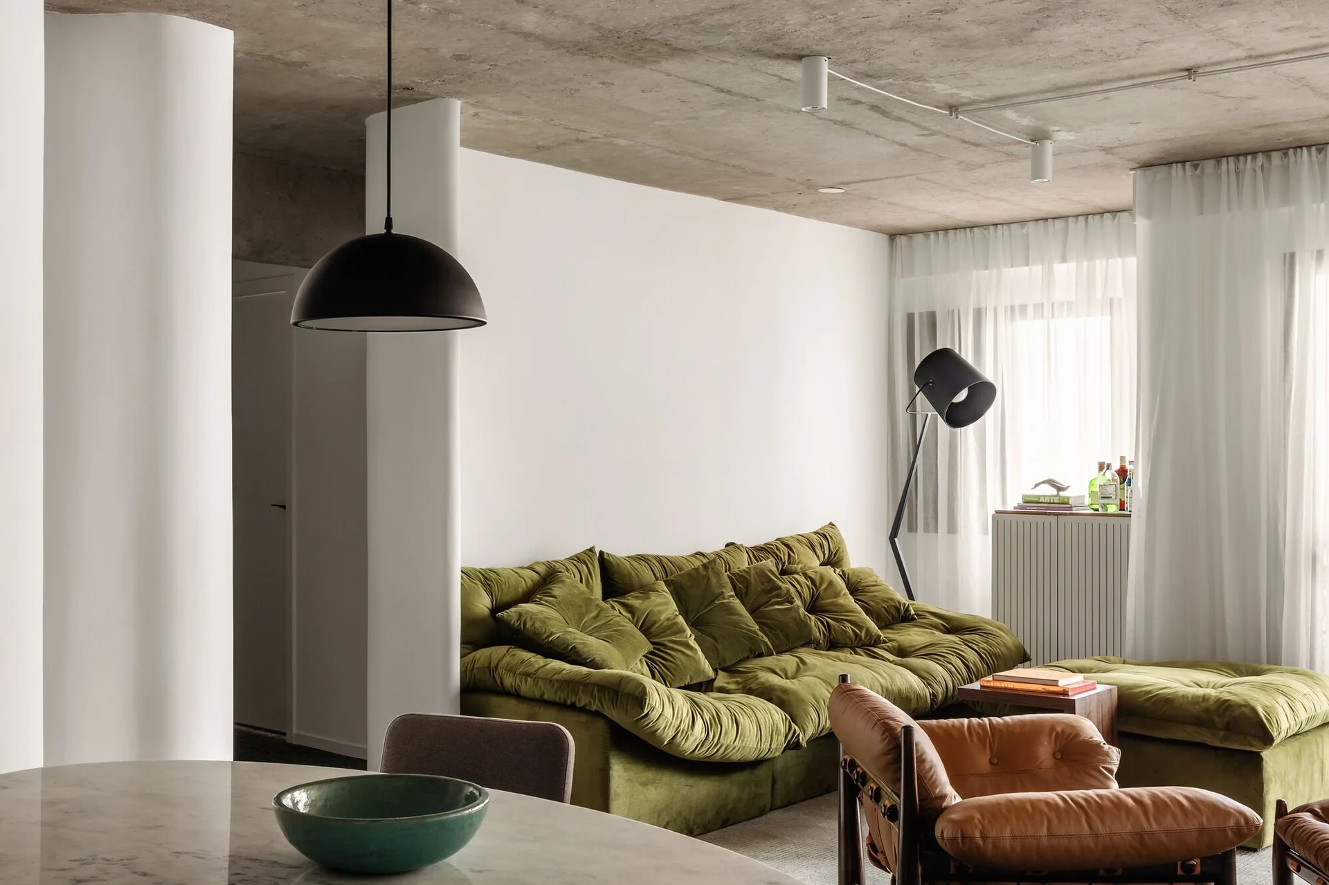 A modern living room features an olive green plush velvet sofa, a tan leather armchair, and an exposed concrete ceiling.