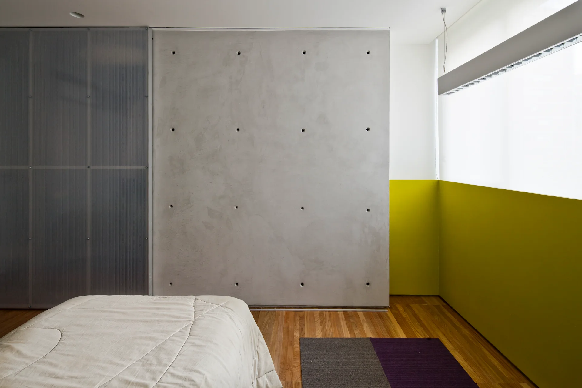 A modern bedroom features an exposed concrete accent wall, a lime green corner wall, a bed with light-colored linens, and natural wood flooring.