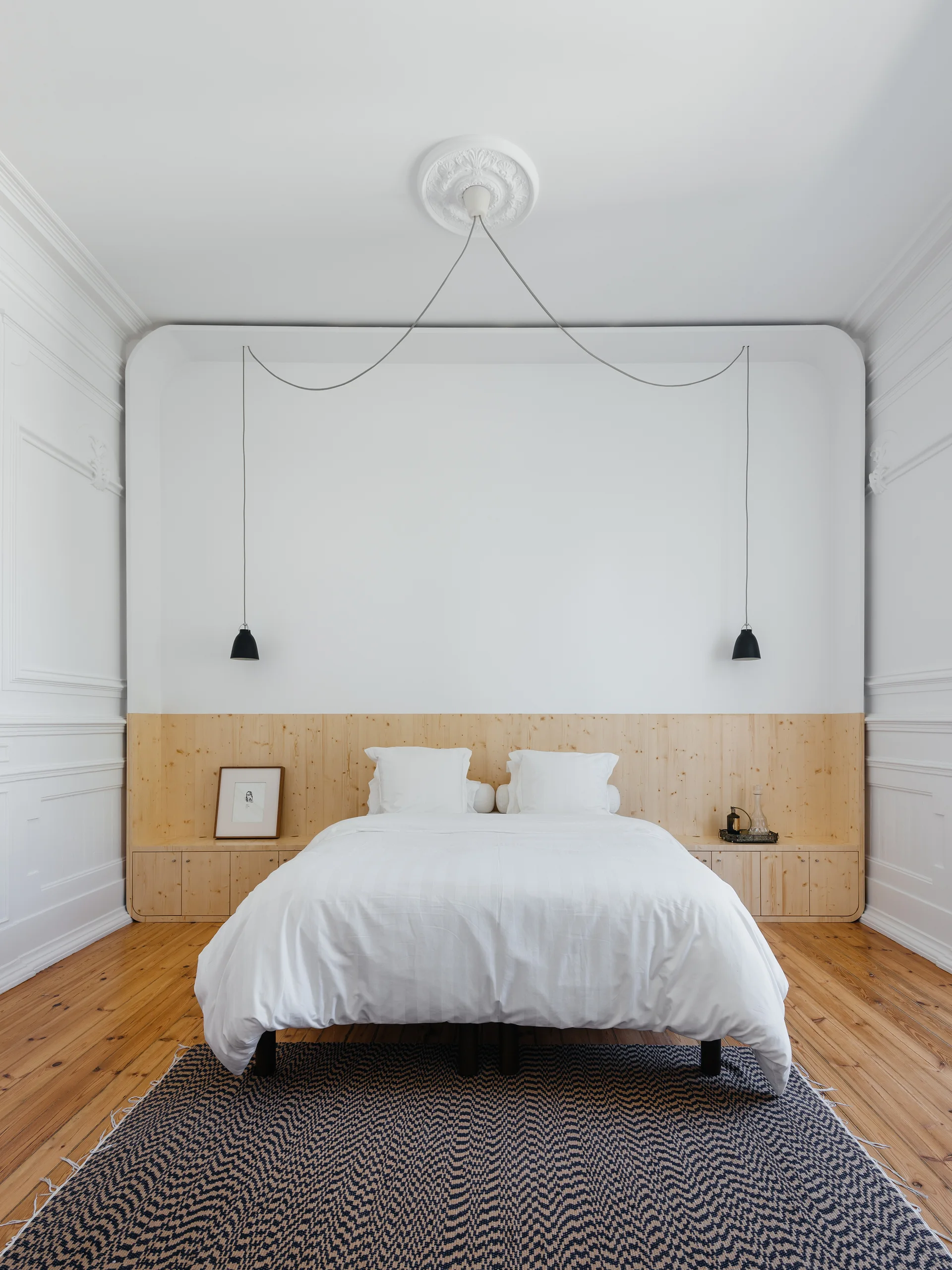 A minimalist white bedroom with a custom pine headboard, two black pendant lights draped from a ceiling medallion, and a dark patterned rug.