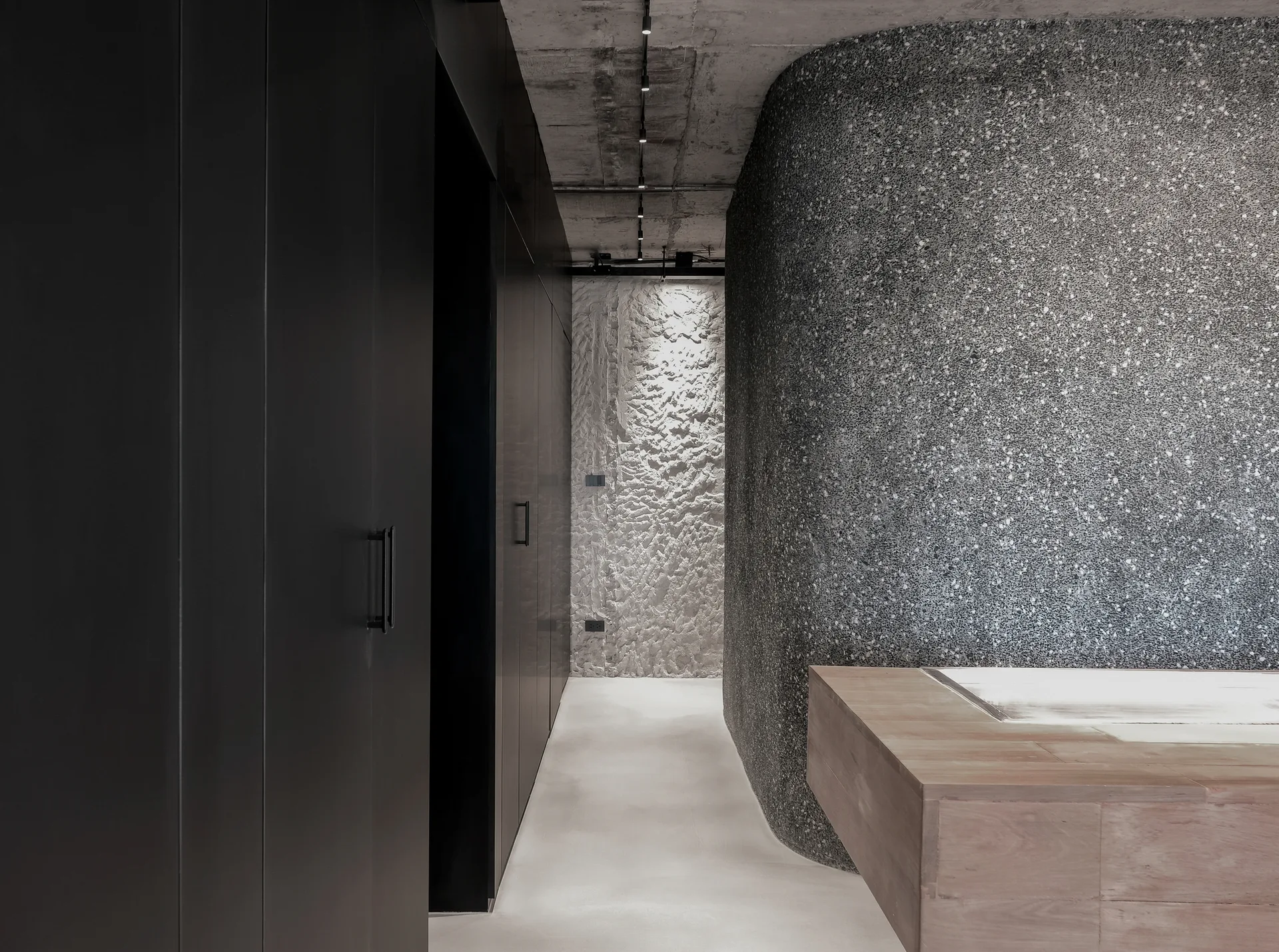 A modern bathroom hallway features a wooden built-in bathtub, a curved terrazzo wall, matte black cabinetry, and an exposed concrete ceiling.