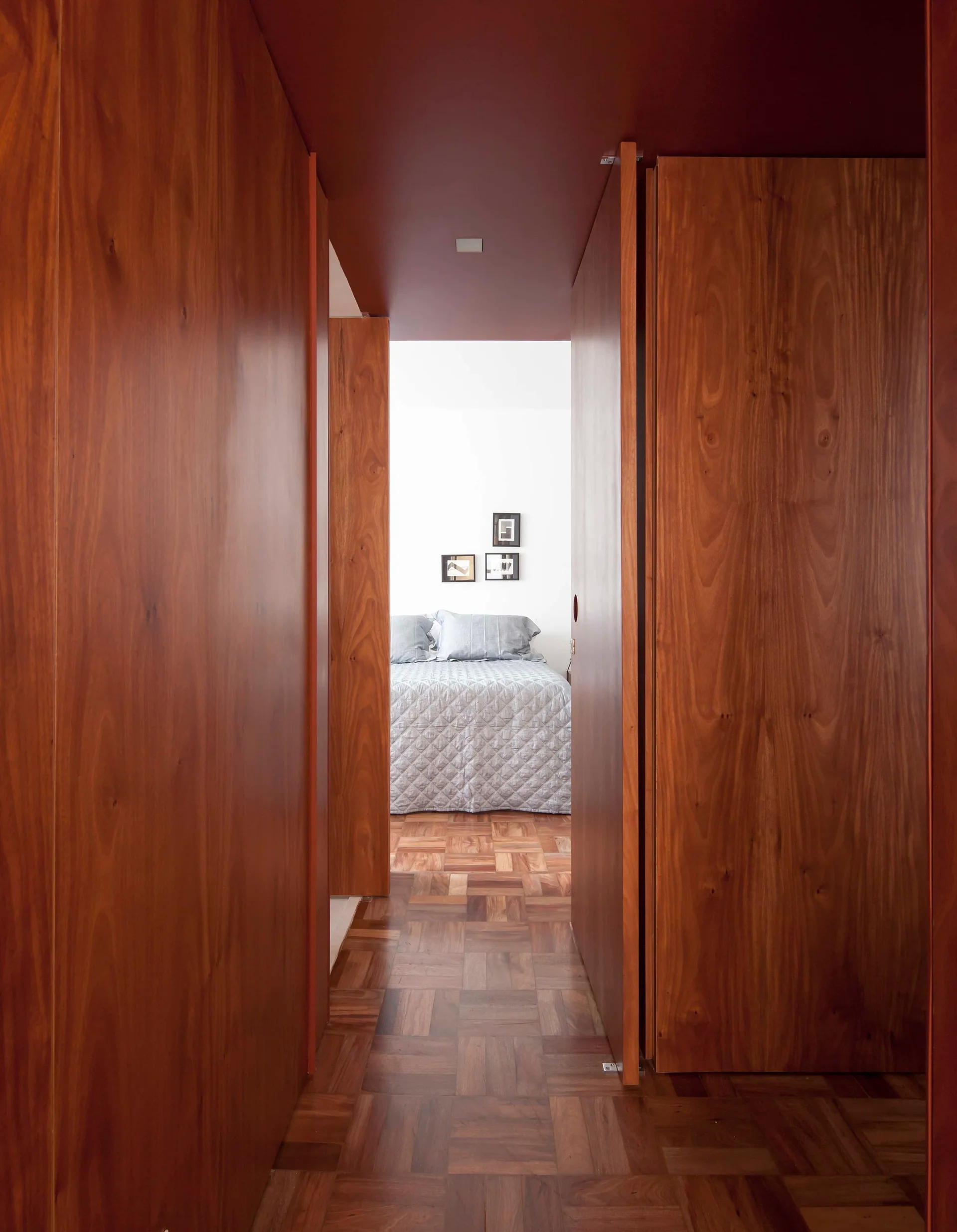 A view down a wood-paneled hallway with a dark red ceiling and parquet floors, looking toward a bright bedroom with a quilted bed.