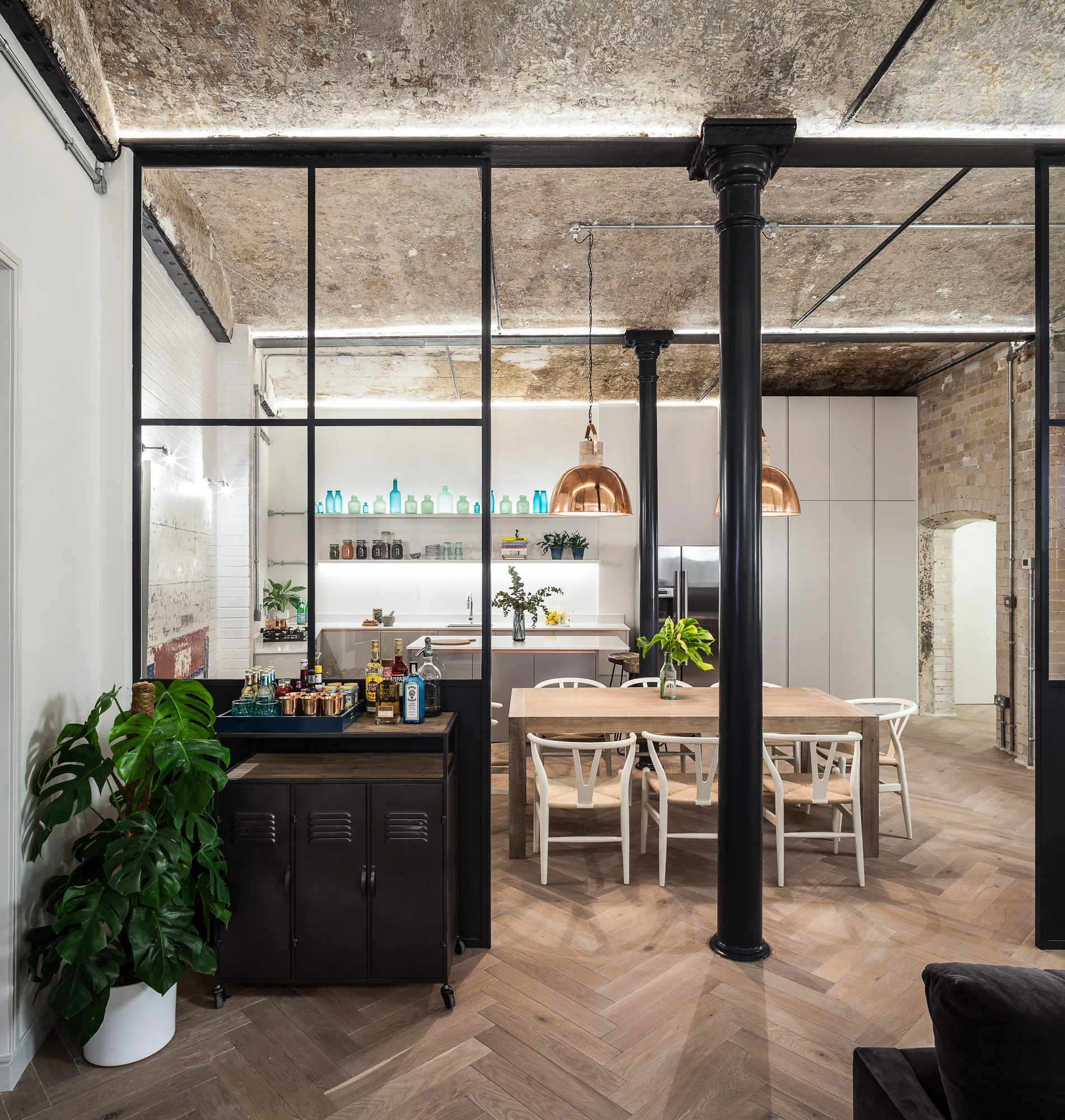 An industrial dining room with a wooden table, white wishbone chairs, copper pendant lights, and a black-framed glass partition wall.