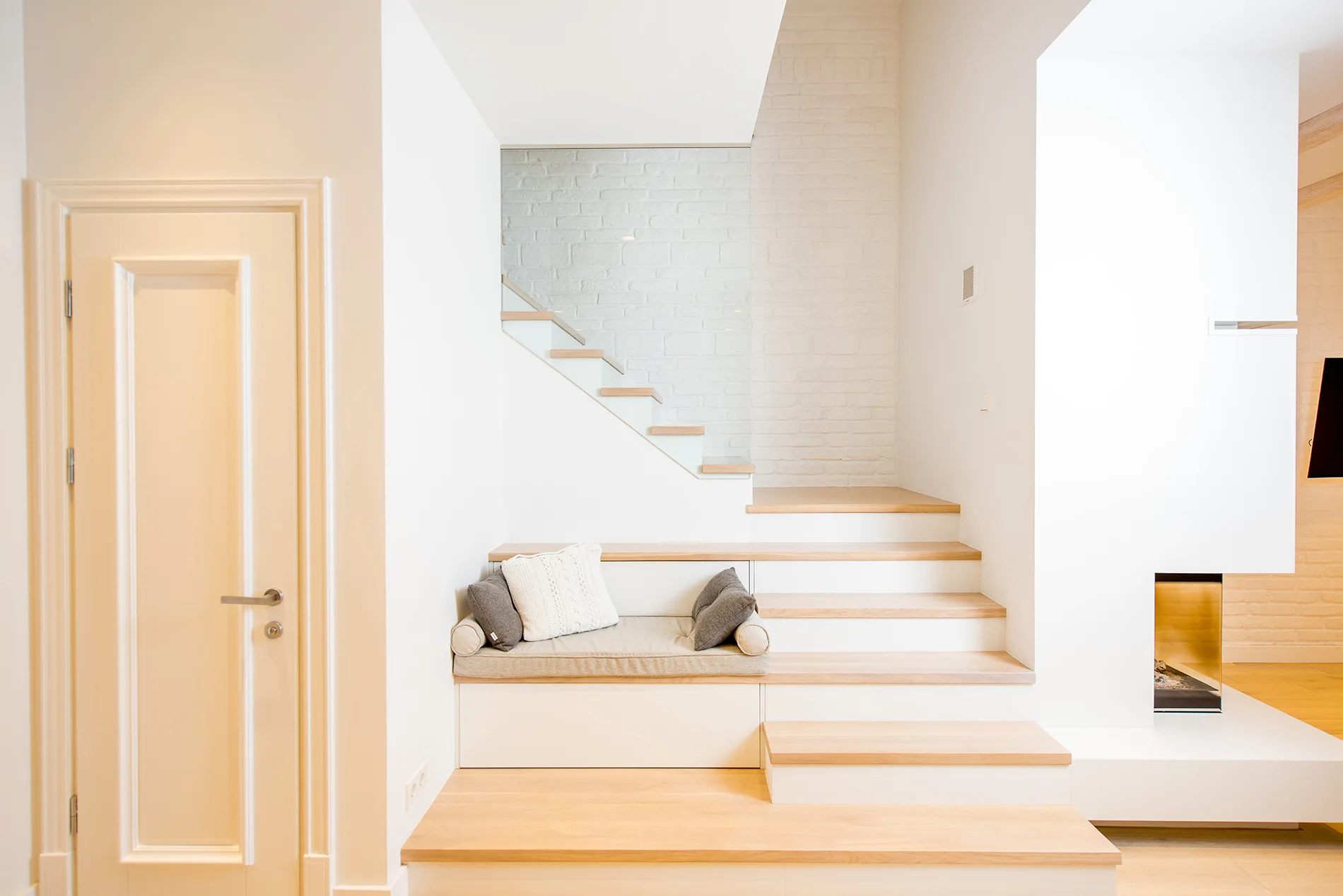 A modern floating wood staircase features an integrated cushioned bench seat with throw pillows against a white painted brick wall.