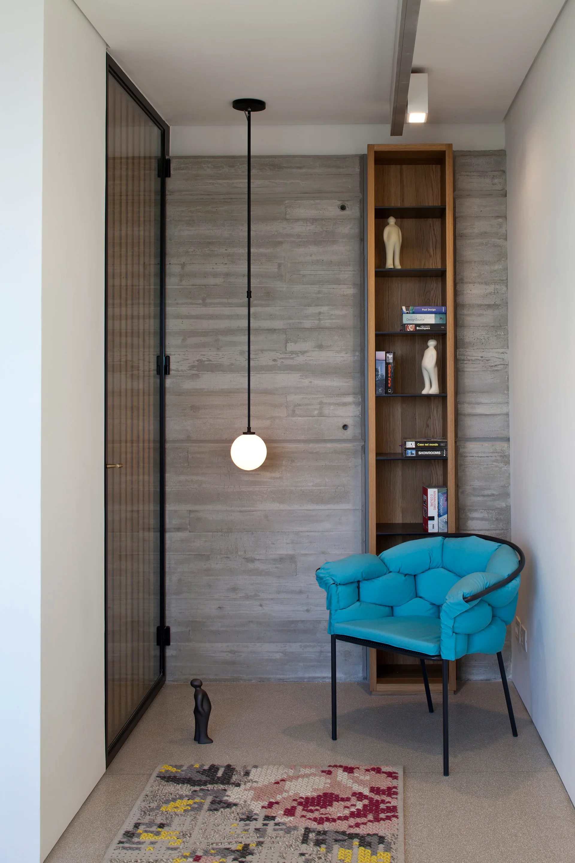 A modern corner nook features a vibrant blue tufted armchair, a tall wooden bookshelf, and a globe pendant light against a concrete wall.