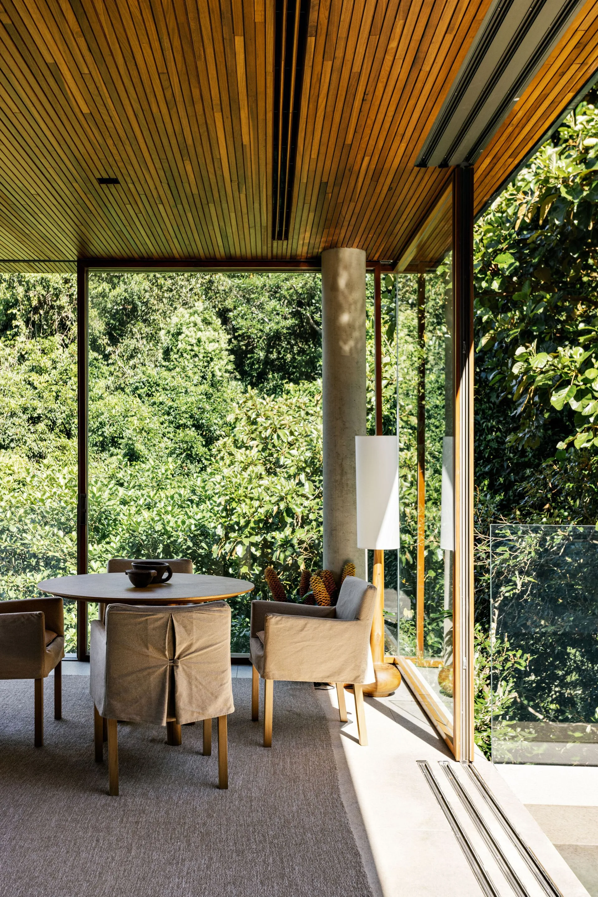 A minimalist dining room featuring a round wooden table, slipcovered chairs, and a slatted wood ceiling next to floor-to-ceiling glass walls.