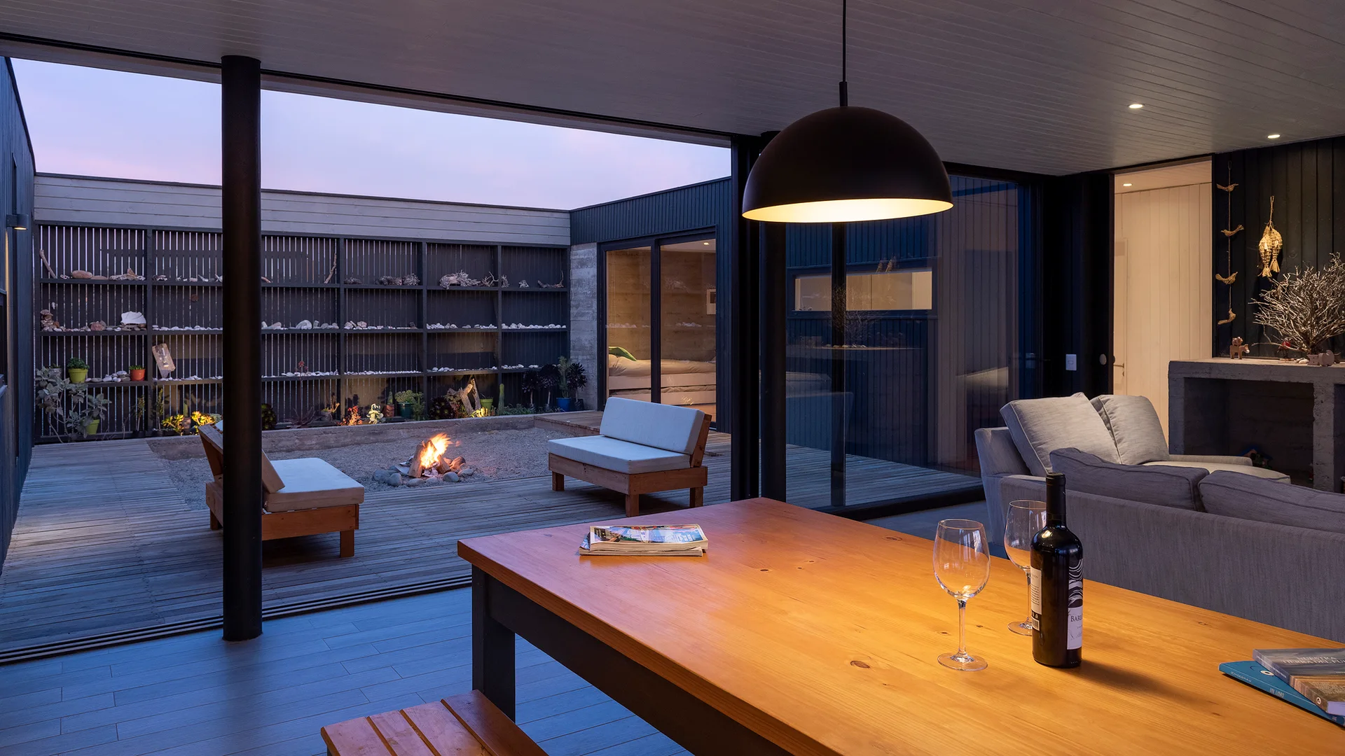 An open-plan dining room with a wooden table looks out onto a decked courtyard featuring a central fire pit and simple wood benches at dusk.