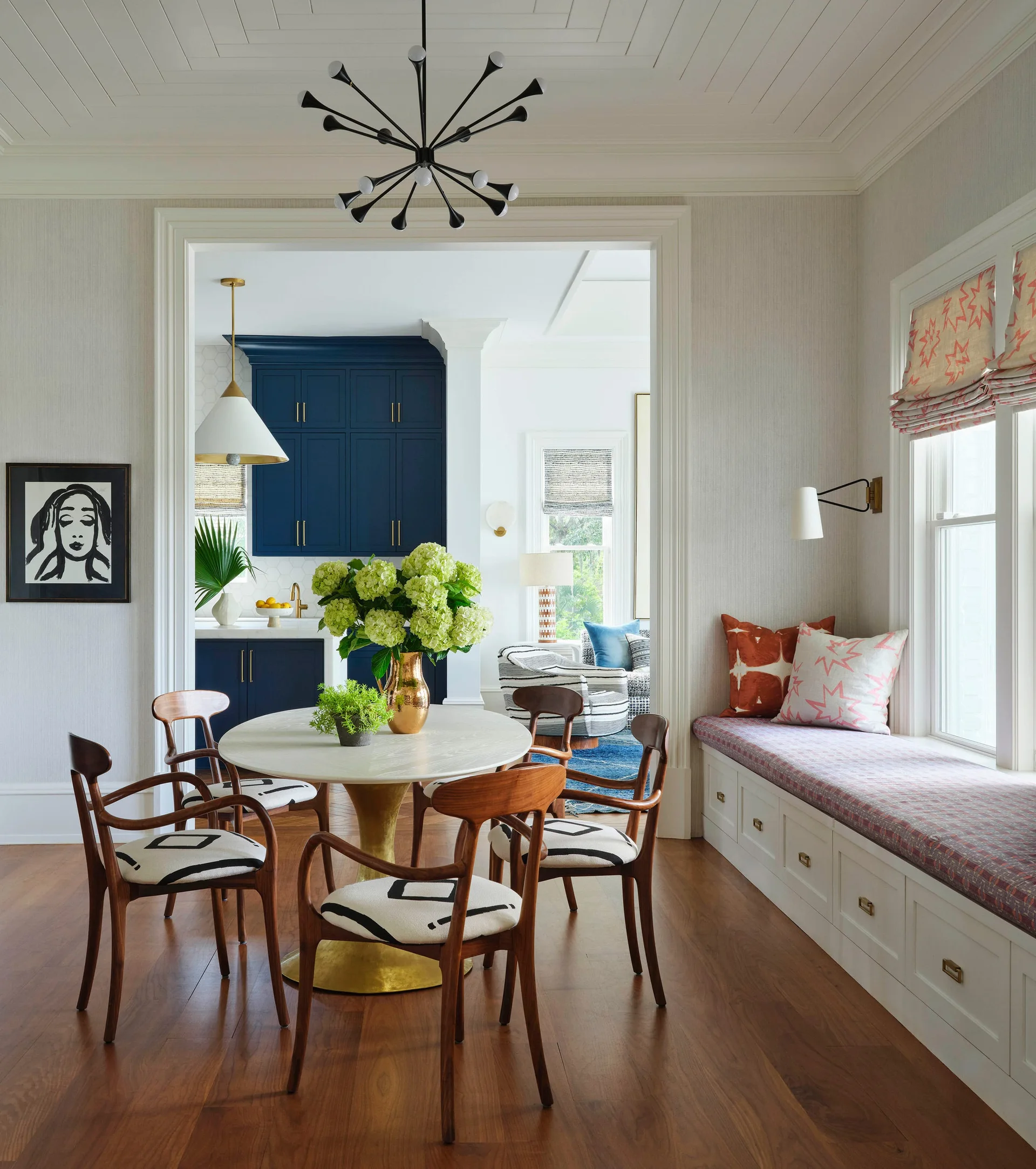 A round dining table with wooden chairs sits near a long white built-in window seat in a dining room leading to a kitchen with navy blue cabinets.