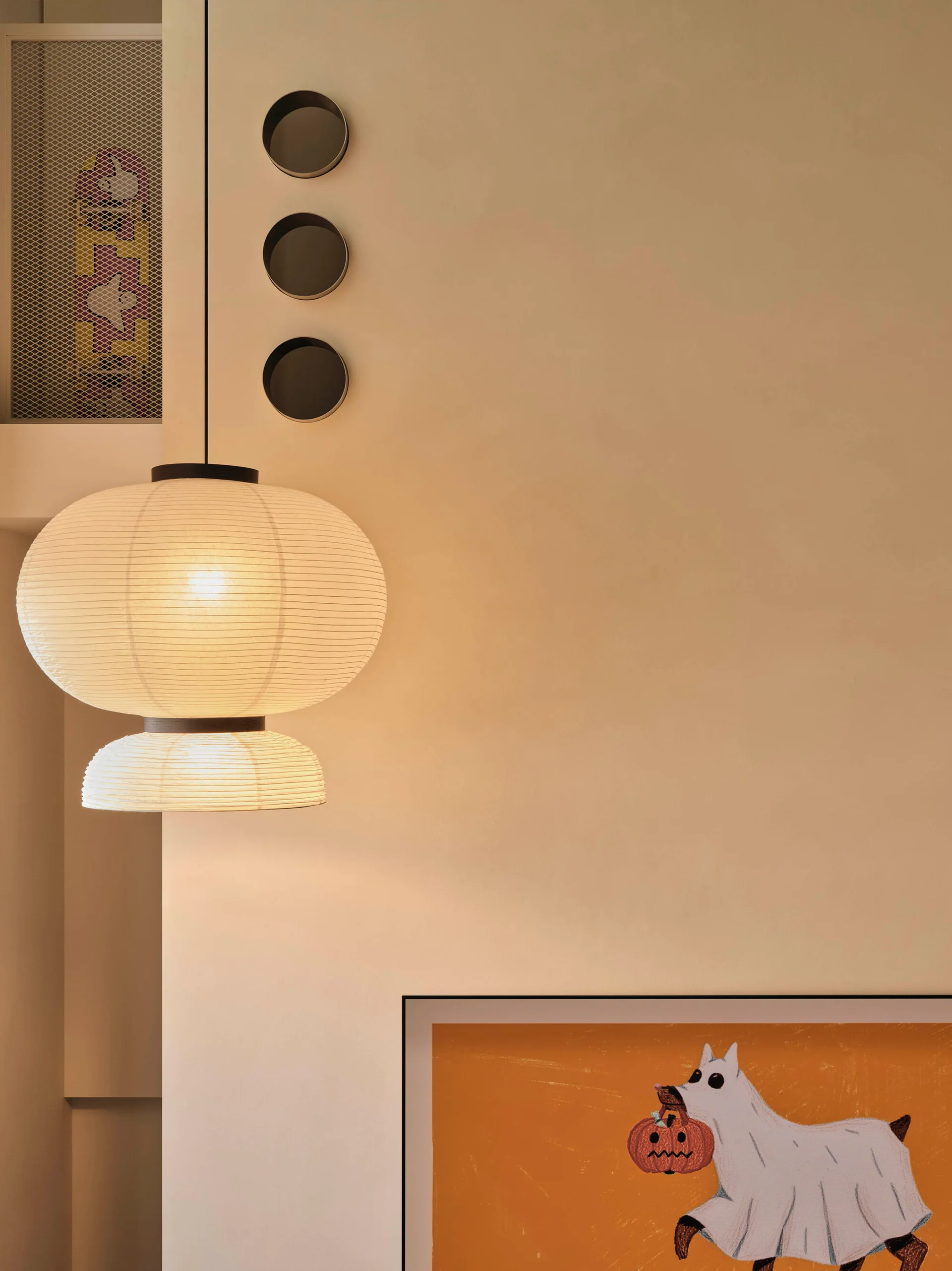 A glowing layered paper pendant light hangs against a cream wall next to three small circular wall decorations and framed orange art.