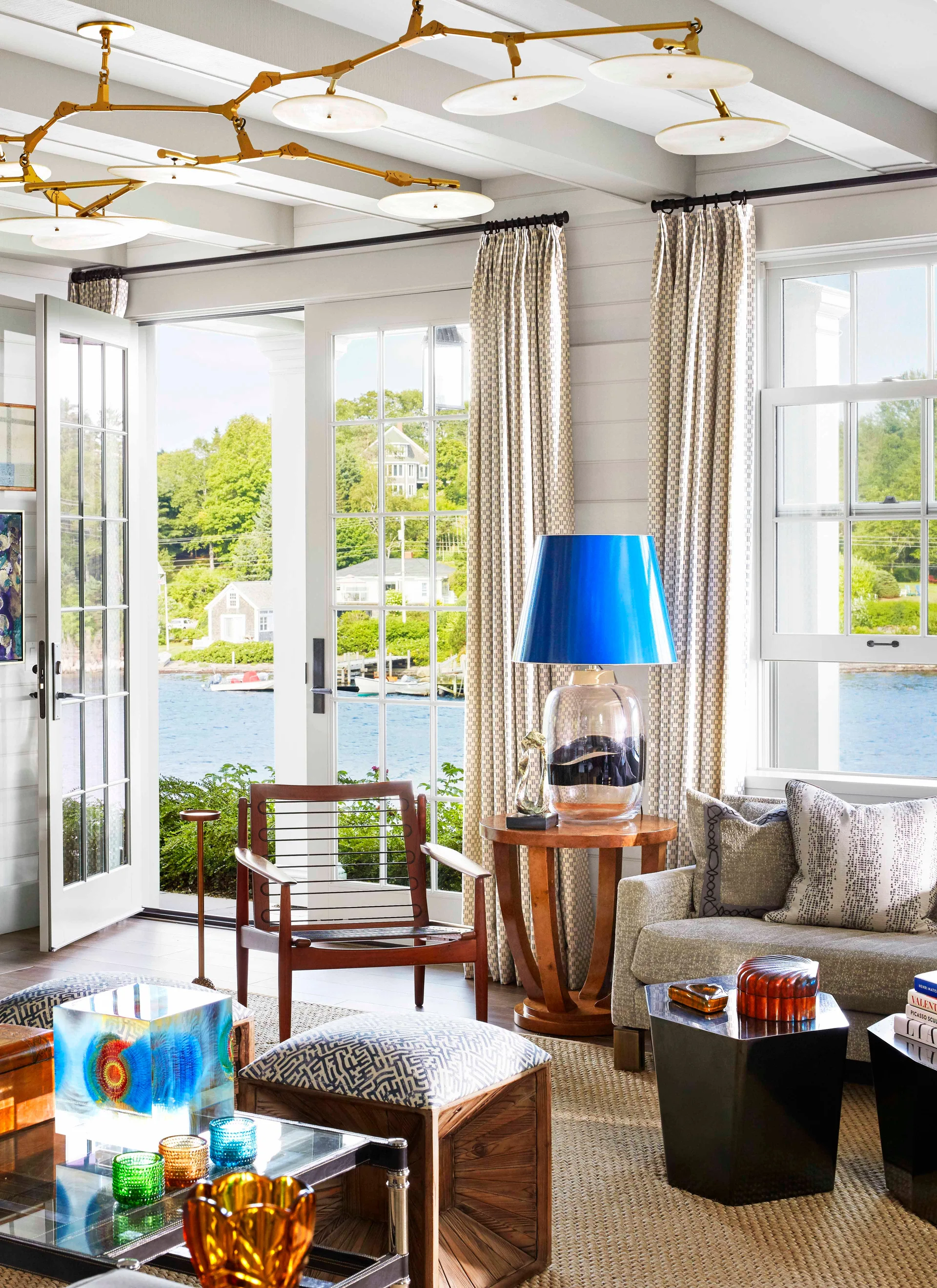 A bright living room features a patterned sofa, a wooden armchair, and a side table with a vibrant blue lamp next to French doors overlooking the water.