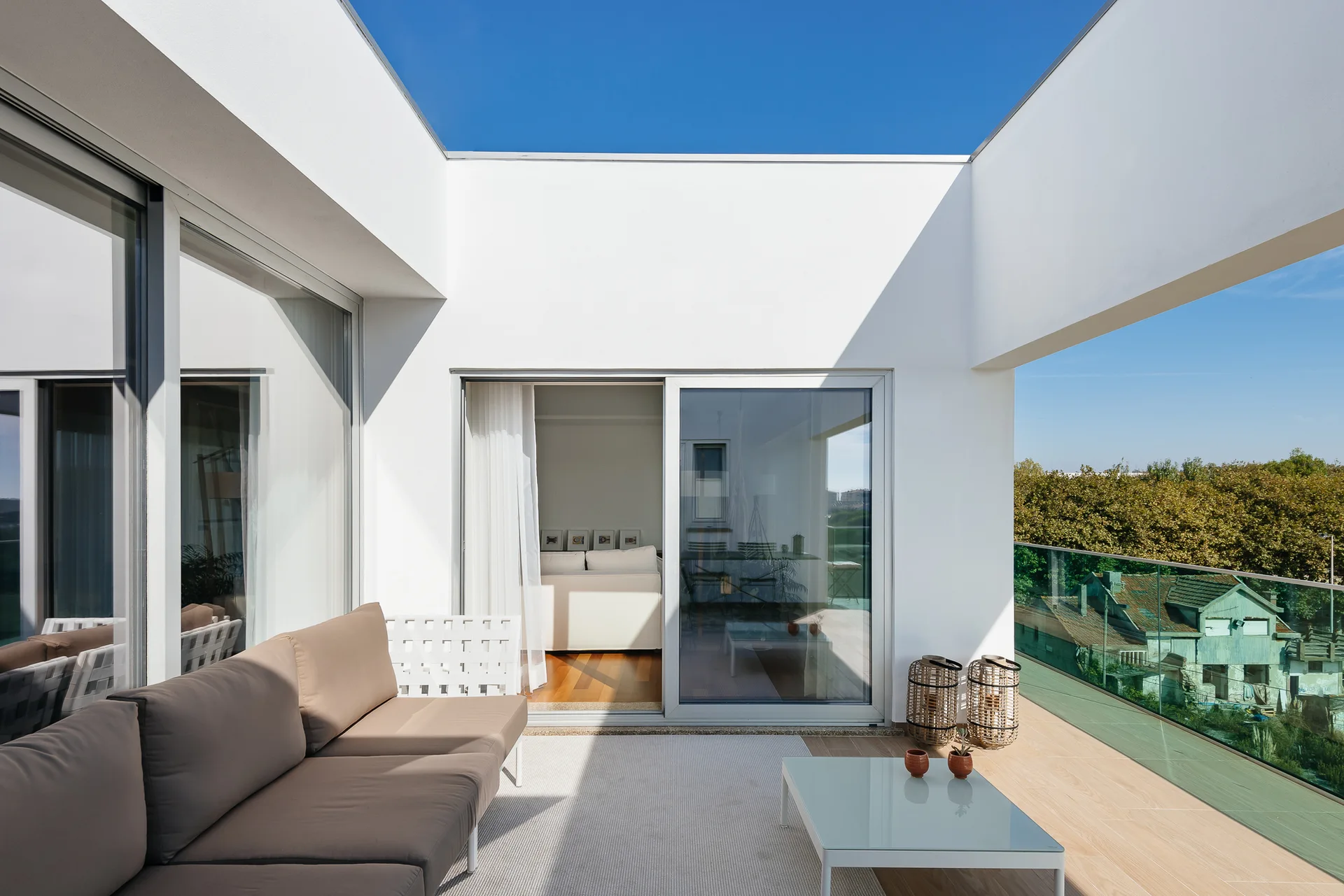 A bright outdoor terrace features a tan sofa, a glass-topped coffee table, and a glass railing overlooking trees.
