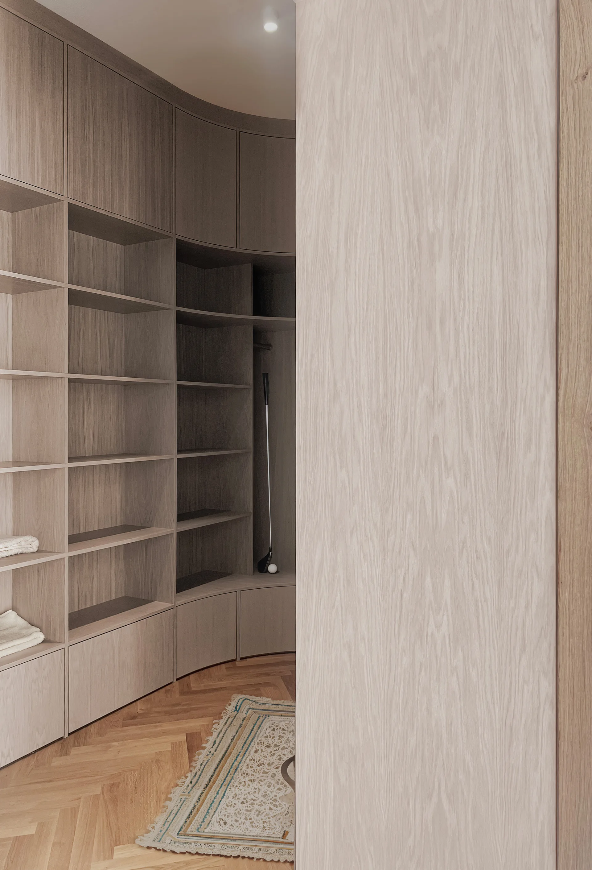 Custom curved light wood shelving units are built along a wall in a dressing room featuring herringbone parquet flooring.