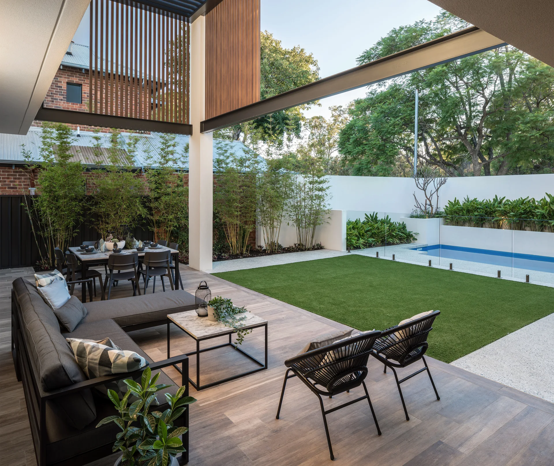 A modern outdoor patio features a grey sectional sofa, black woven chairs, a dining set, and a green lawn next to a fenced swimming pool.