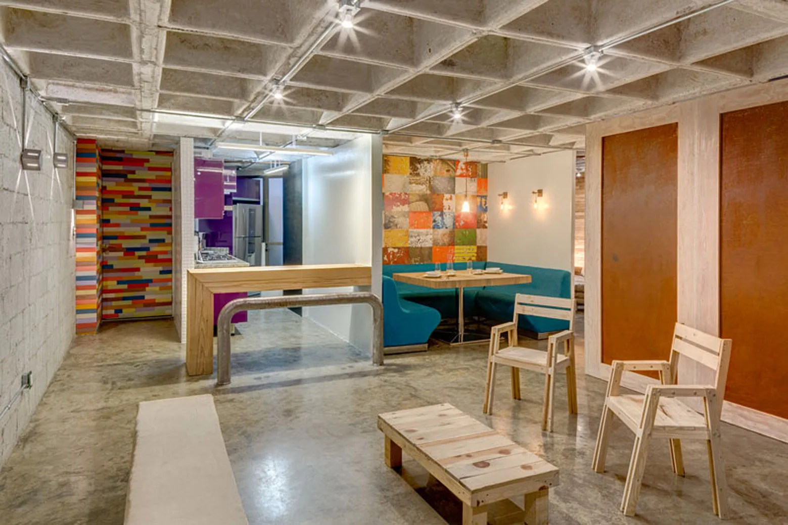 Industrial dining area featuring a teal corner booth, a wooden table, and simple wooden chairs under an exposed concrete waffle slab ceiling.