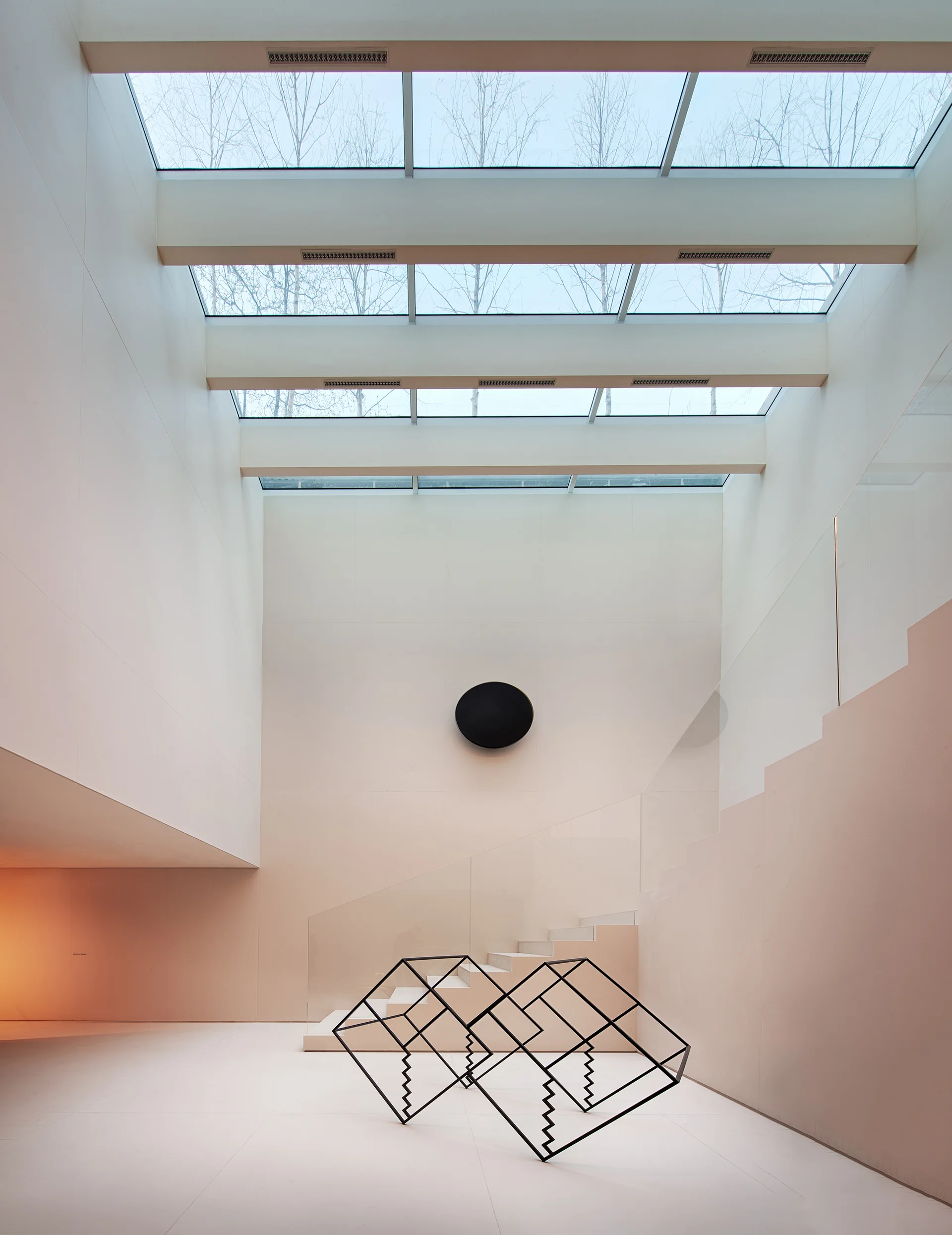 A minimalist double-height atrium features a pale geometric staircase with a glass railing, a black wireframe floor sculpture, and overhead skylights.