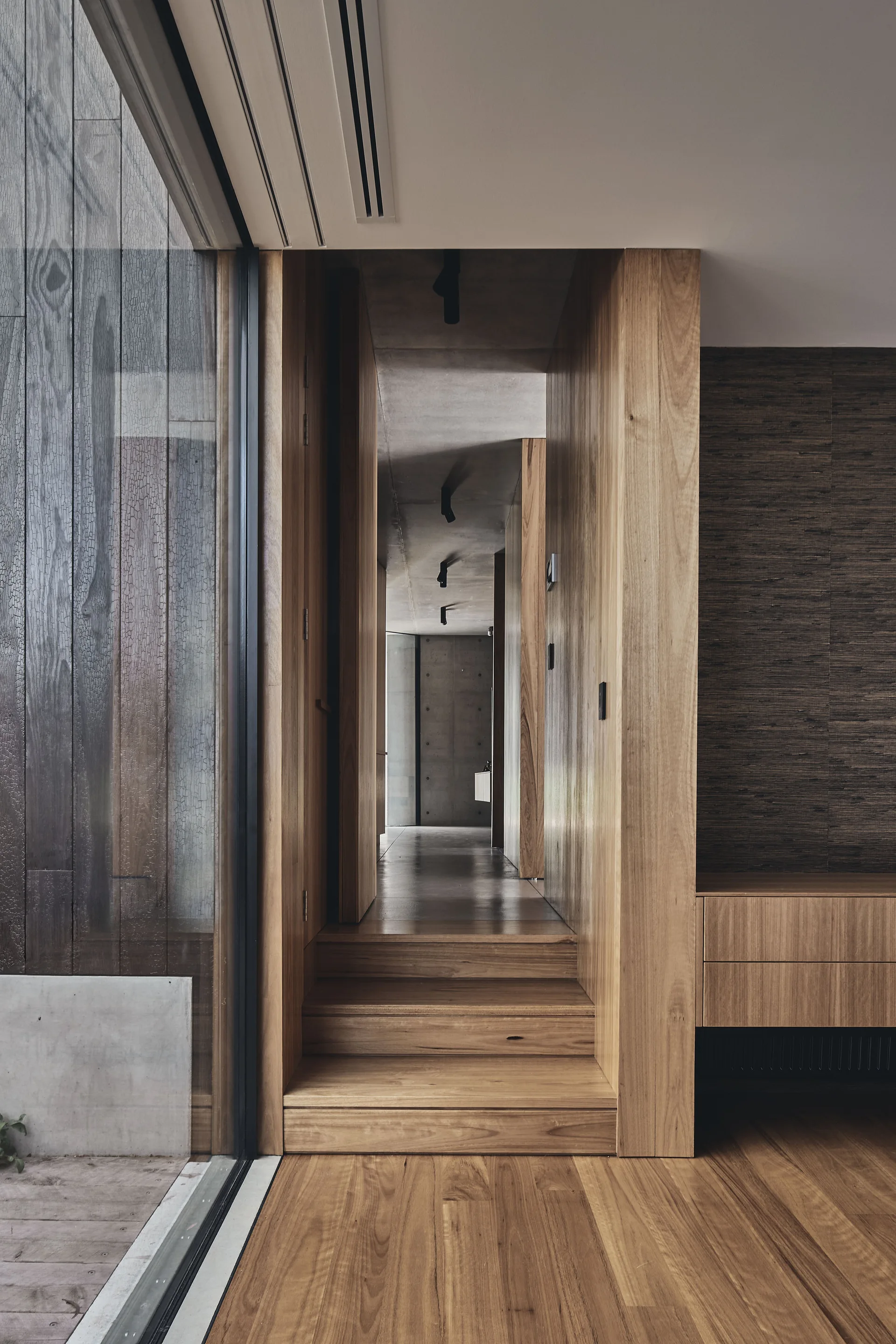 A modern hallway features wooden steps, matching timber-paneled walls and floors, and an industrial exposed concrete ceiling with track lighting.