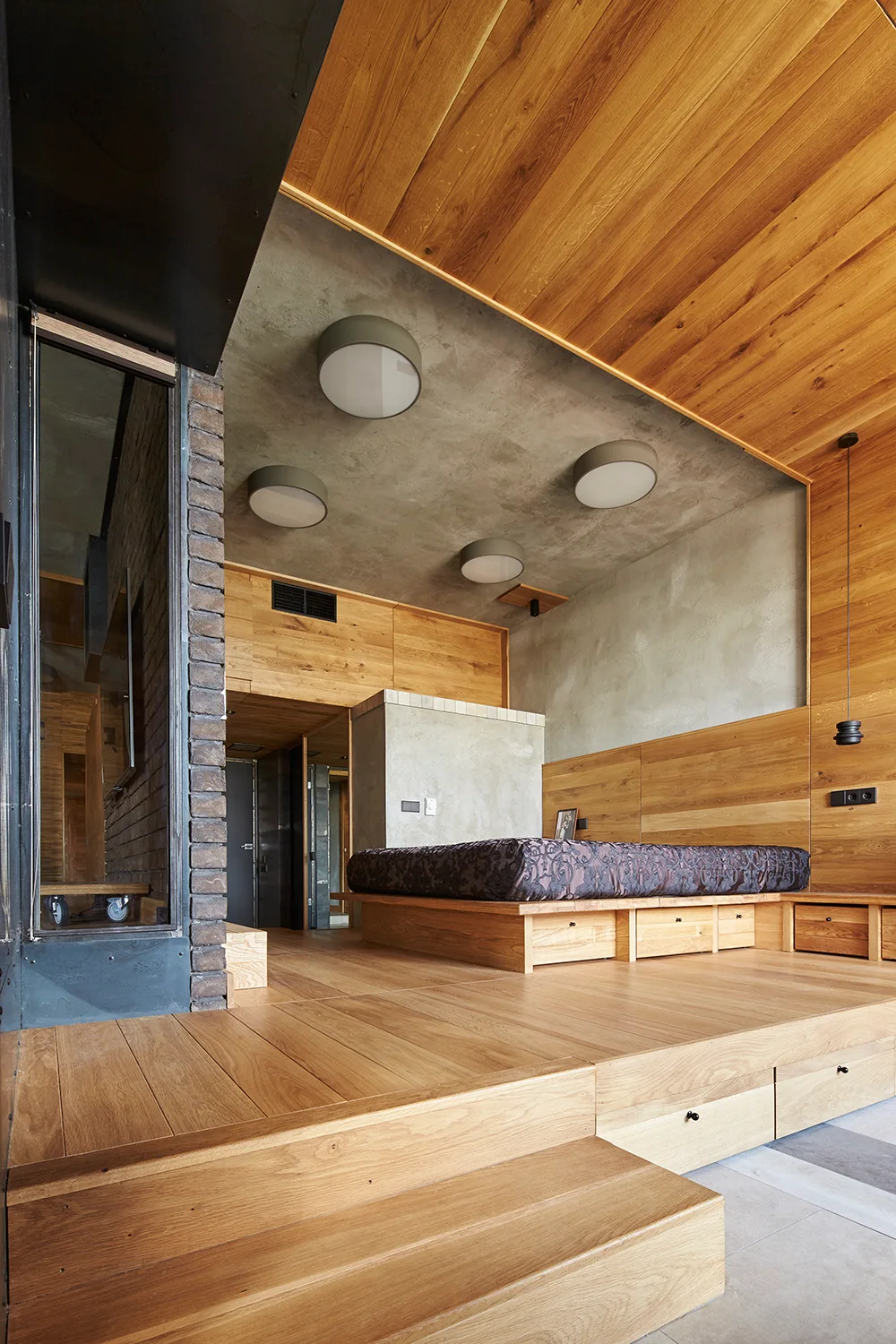 A minimalist bedroom features a raised wooden platform bed with built-in drawers, wood-paneled walls, and a recessed raw concrete ceiling.