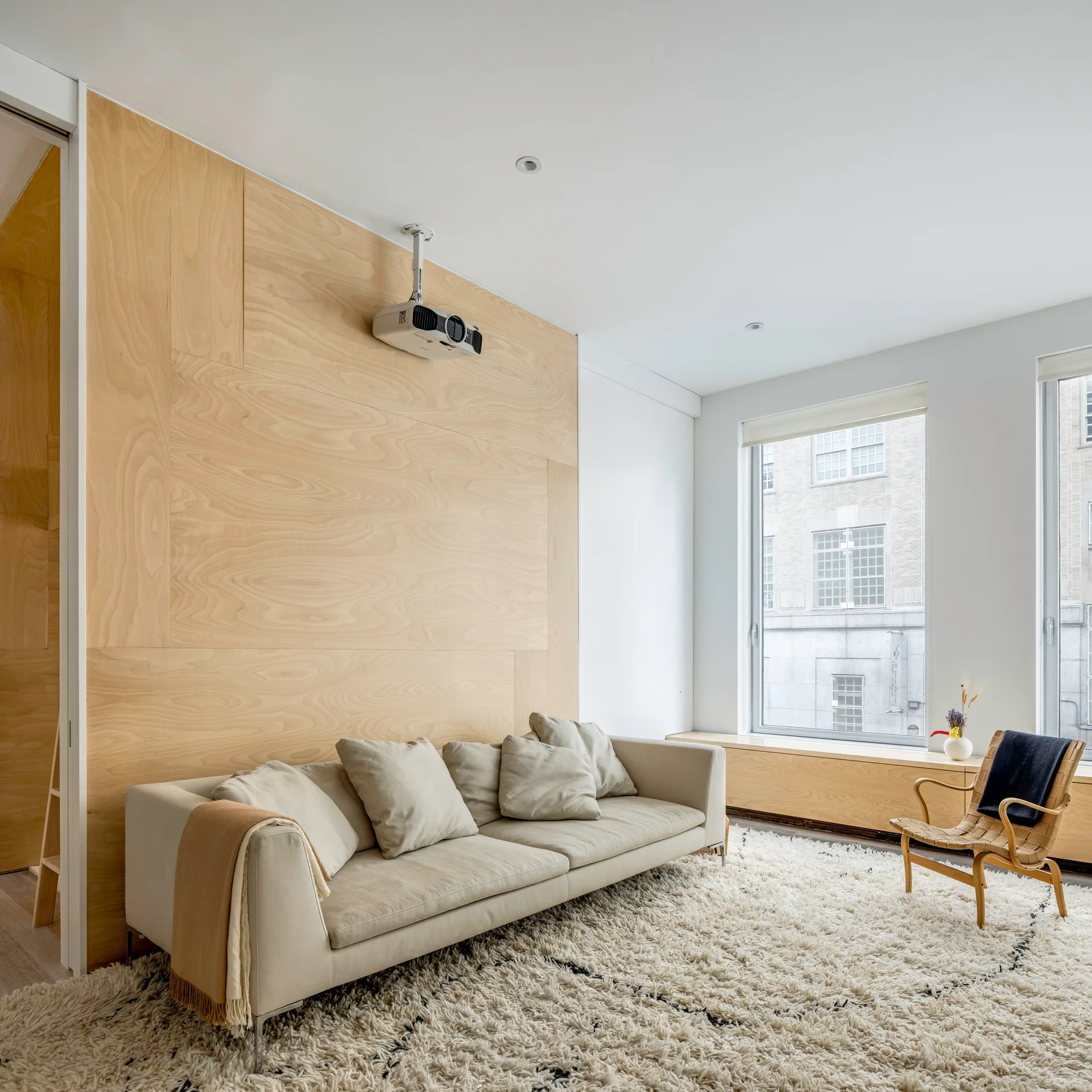 A minimalist living room featuring a cream sofa, a plush shag rug, and a light-toned plywood paneled feature wall under a white ceiling.