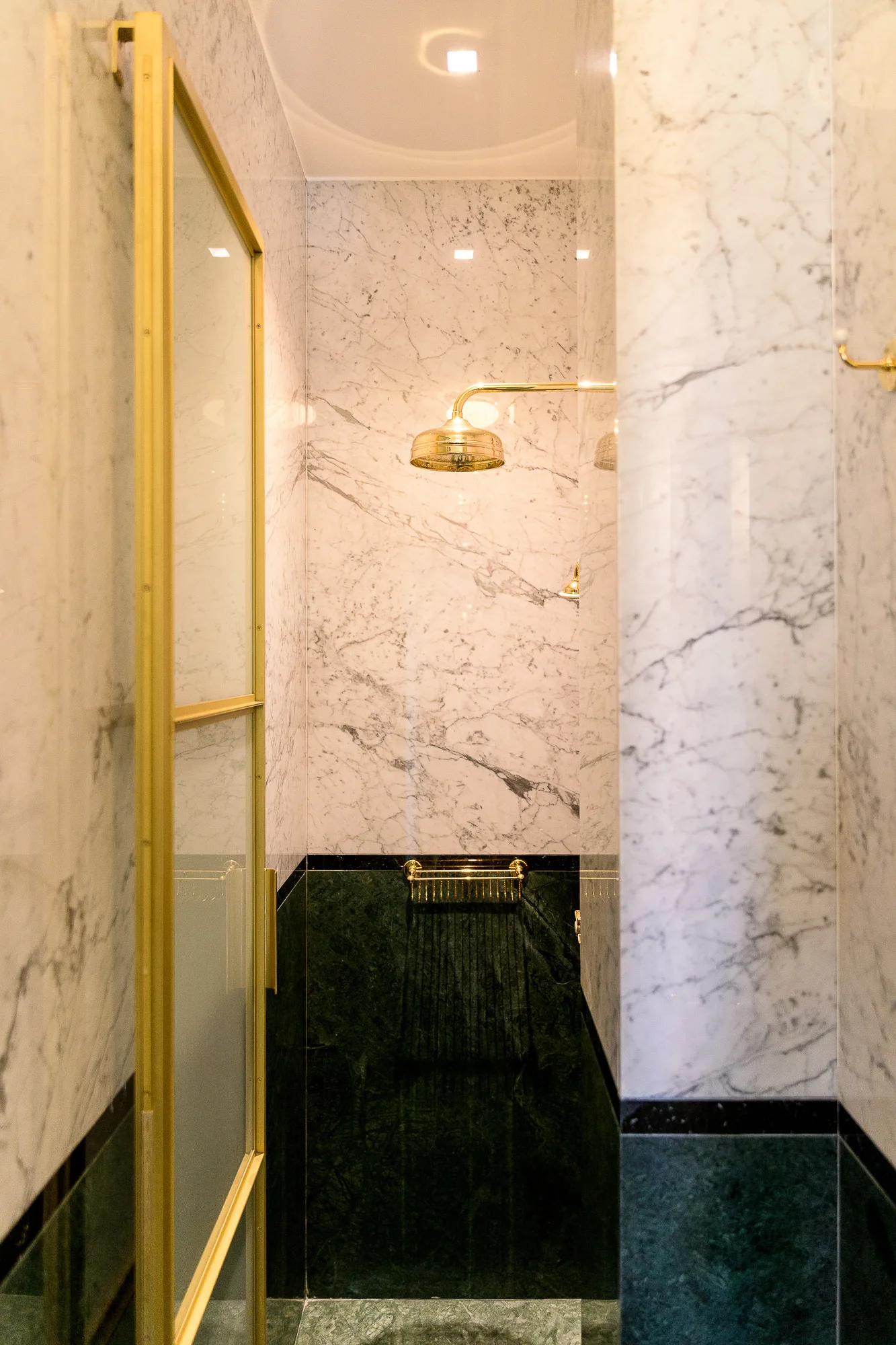 A walk-in shower features white veined marble walls, a dark green stone base, and polished gold fixtures including a round shower head and metal frame.