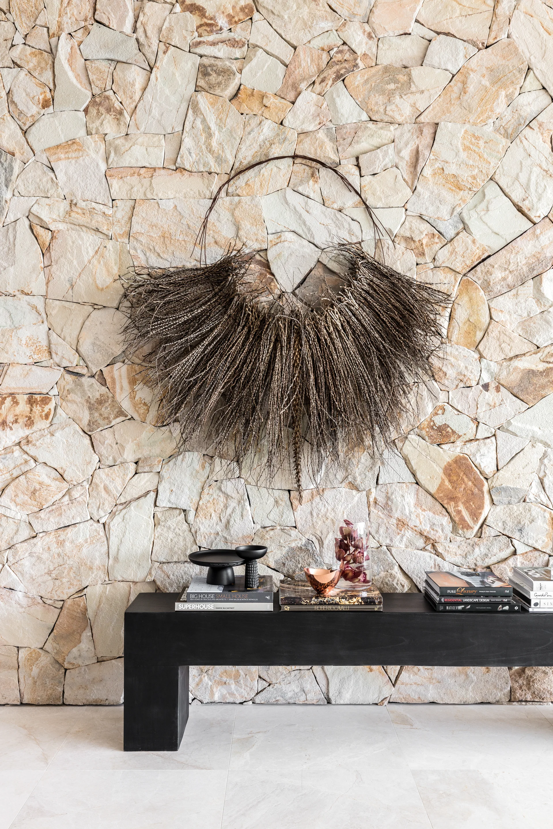 A black wooden console table with stacked books sits against a rustic beige stone wall decorated with a circular grass sculpture.