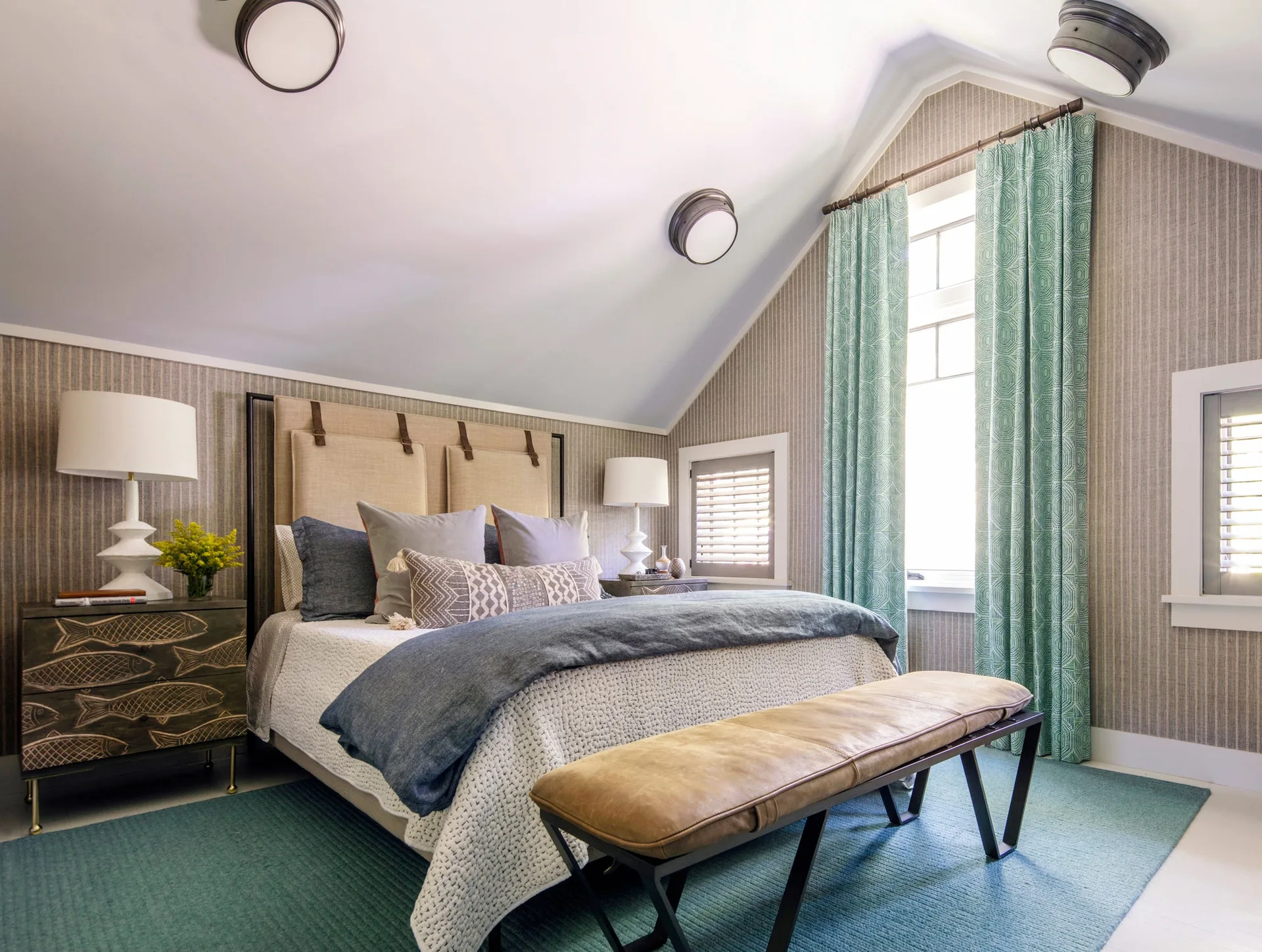 An attic bedroom features an upholstered headboard, a leather bench, striped wallpaper, and patterned teal curtains framing a window.