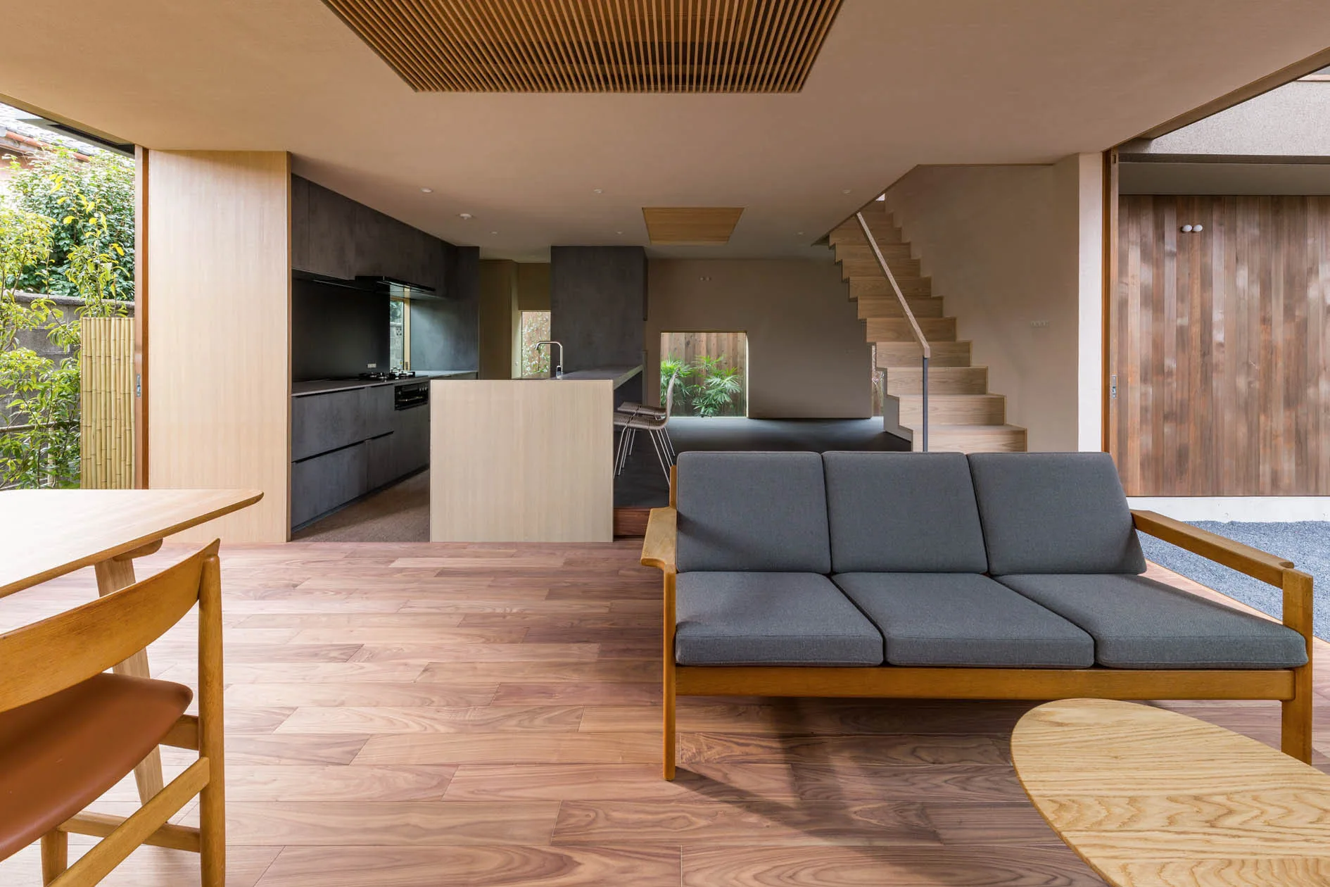 A modern open-plan living room with a grey wood-frame sofa, warm wood flooring, a matching staircase, and dark kitchen cabinets in the background.