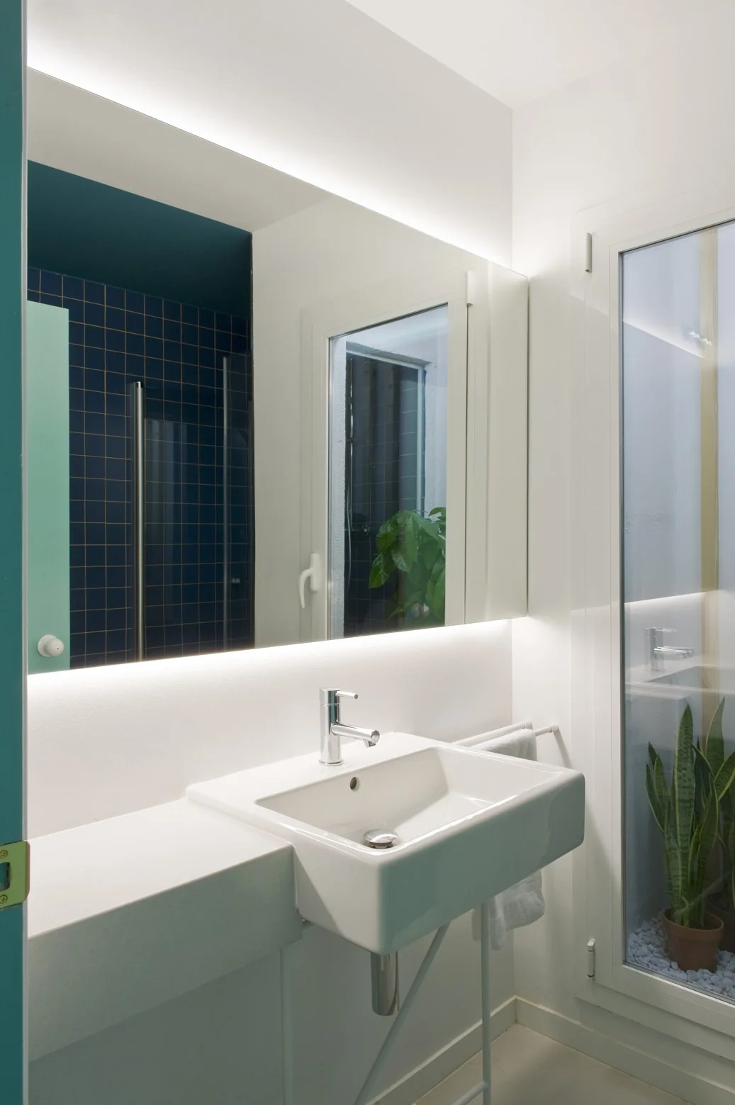 A modern bathroom features a white rectangular sink with chrome faucet, a large backlit mirror, and a window showing a potted plant in a lightwell.