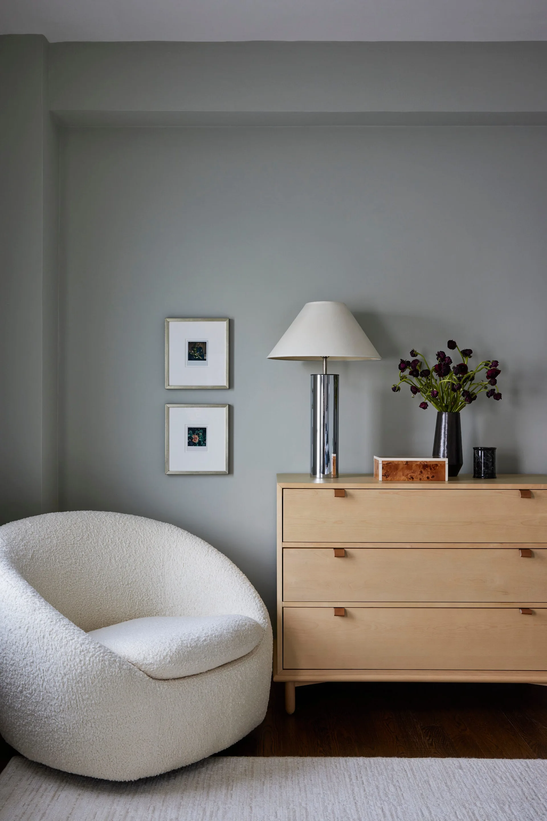 A bedroom corner features a curved white bouclé chair next to a light wood dresser on a dark wood floor.