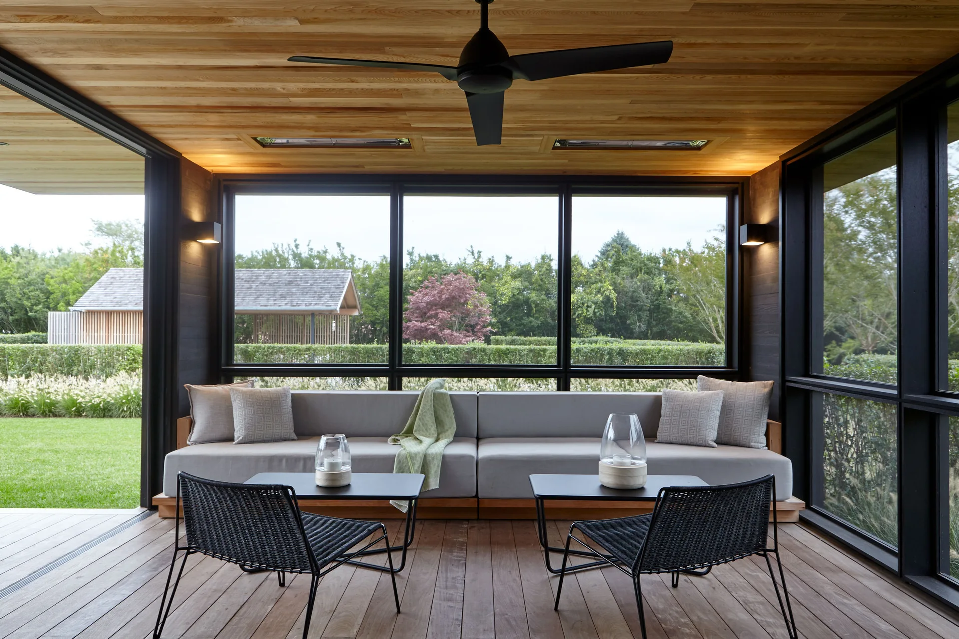 A modern covered porch features a long grey sofa, two black woven chairs, a natural wood ceiling with a black ceiling fan, and large windows.