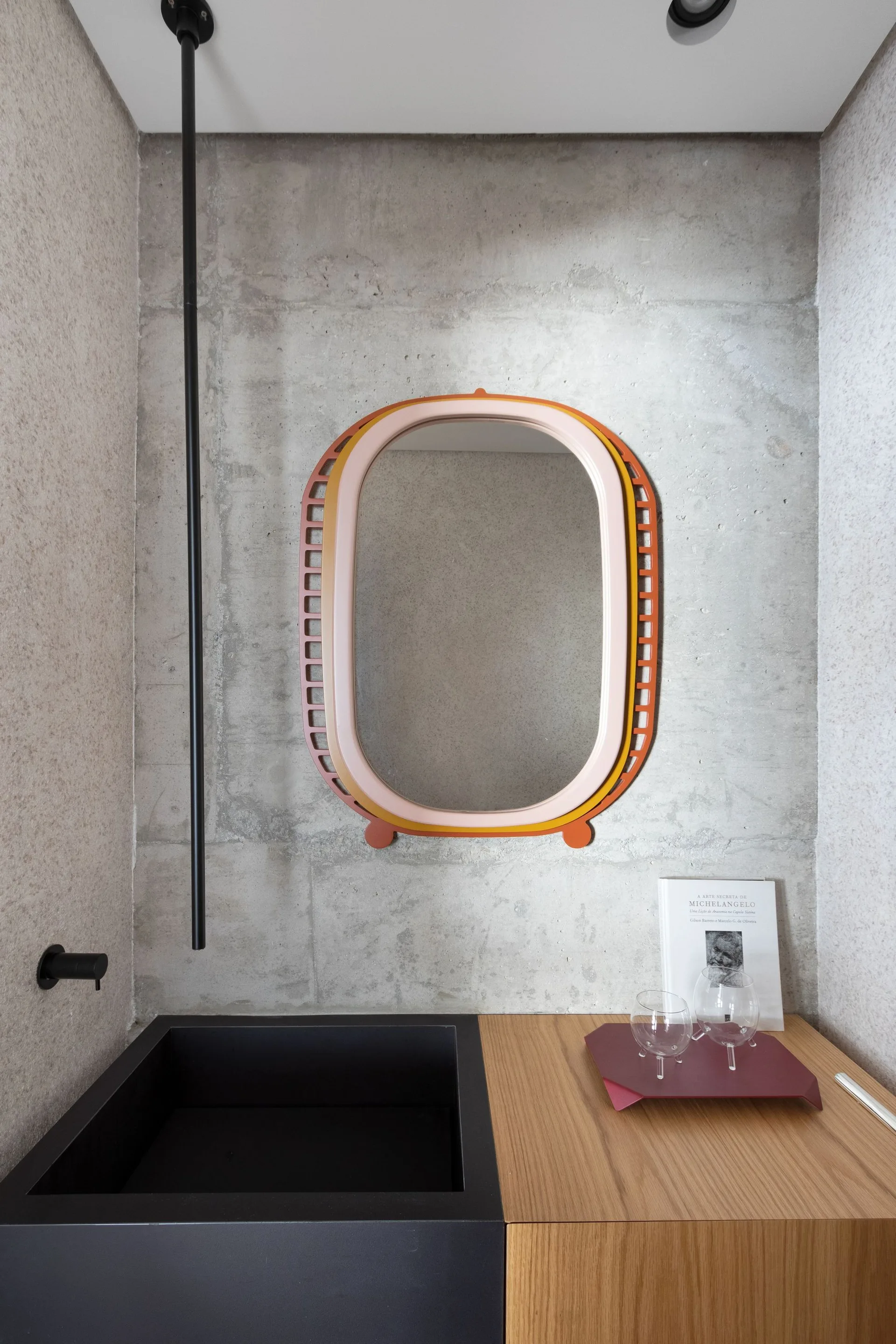 A modern bathroom vanity featuring a black sink and wood cabinet against a textured grey concrete wall with a colorful layered oval mirror.