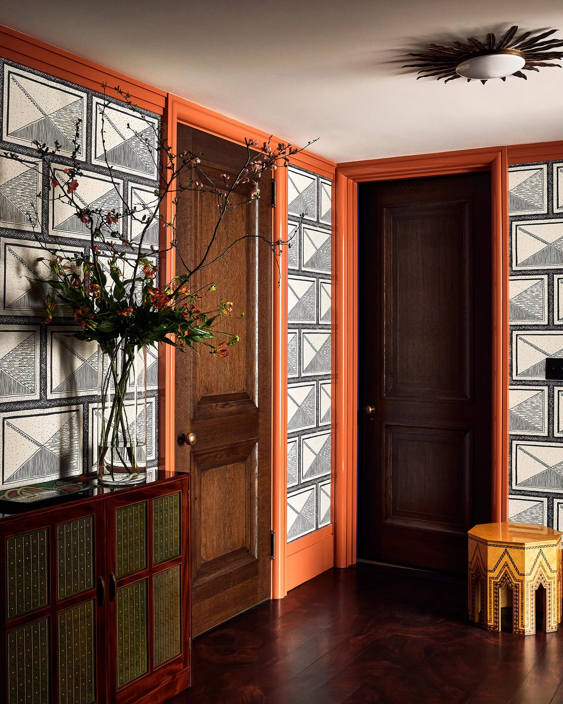 A hallway with geometric black and white wallpaper, vibrant orange painted door trim, dark wood doors, a wood console table, and dark floors.