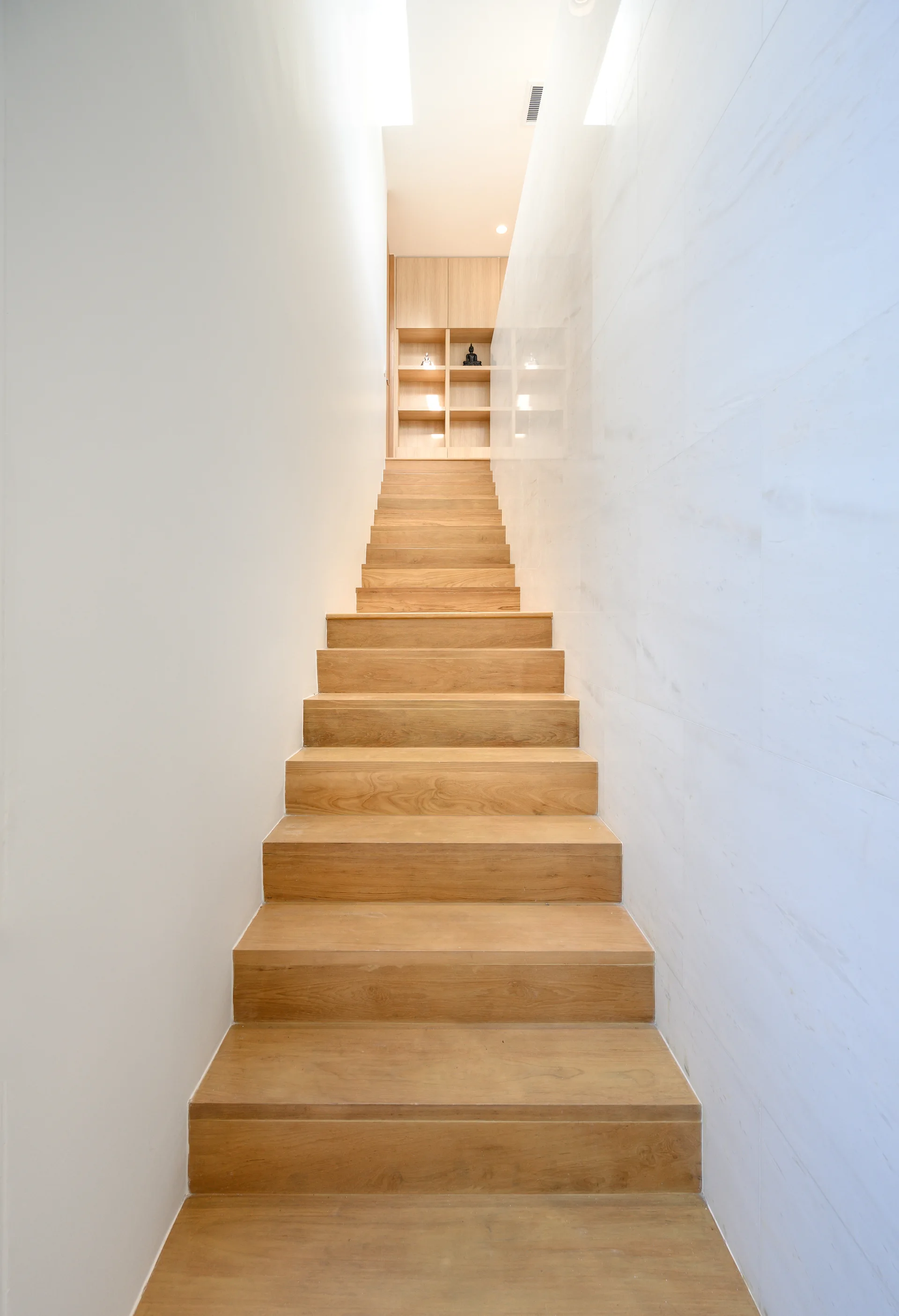 A minimalist indoor staircase with light wood treads is flanked by a white wall and a stone-tiled wall, leading up to built-in shelving.