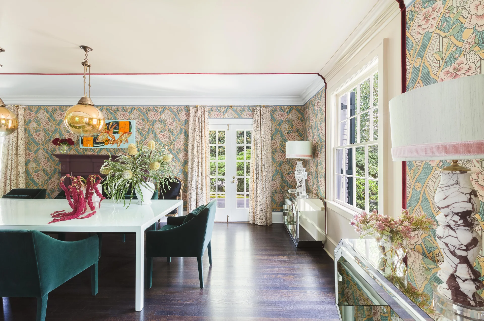 A dining room features a white table surrounded by emerald green velvet chairs, set against ornate floral wallpaper and dark wood floors.