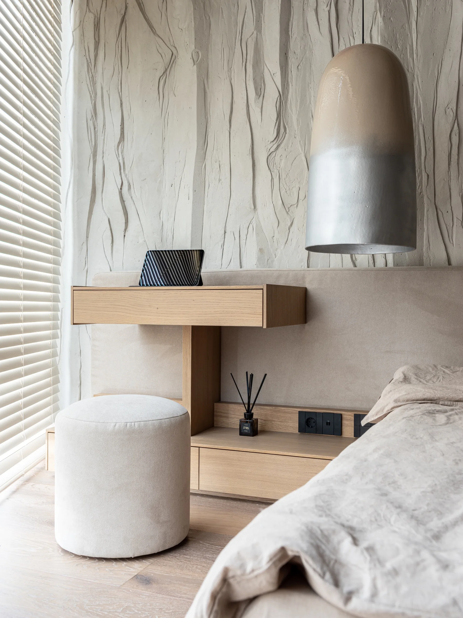 A modern bedroom corner featuring a floating wooden bedside desk, a round fabric stool, and a highly textured plaster wall with a pendant light.