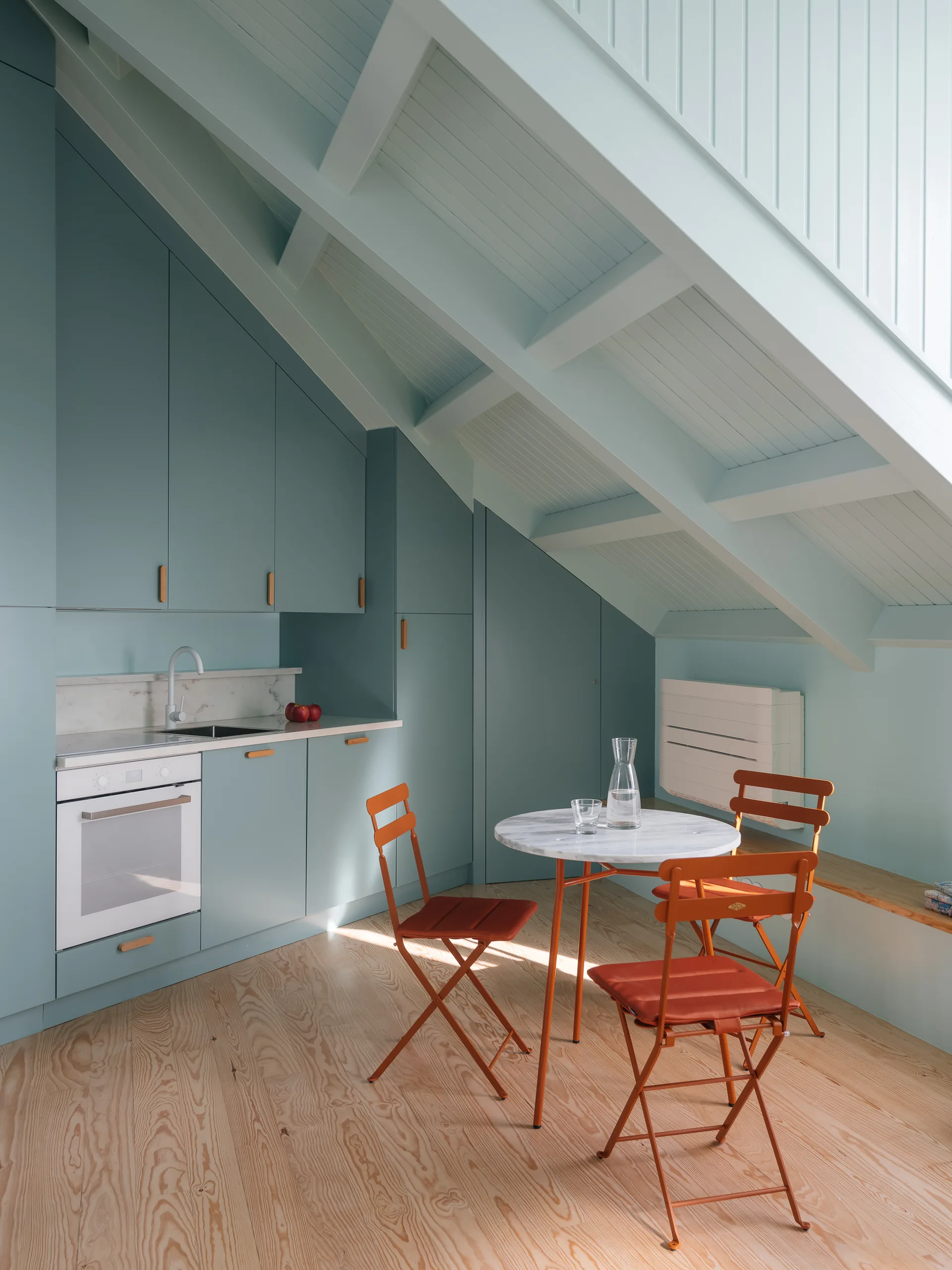 A compact attic kitchenette features blue-green built-in cabinets under a sloped ceiling, a marble bistro table, and orange folding chairs on wood floors.