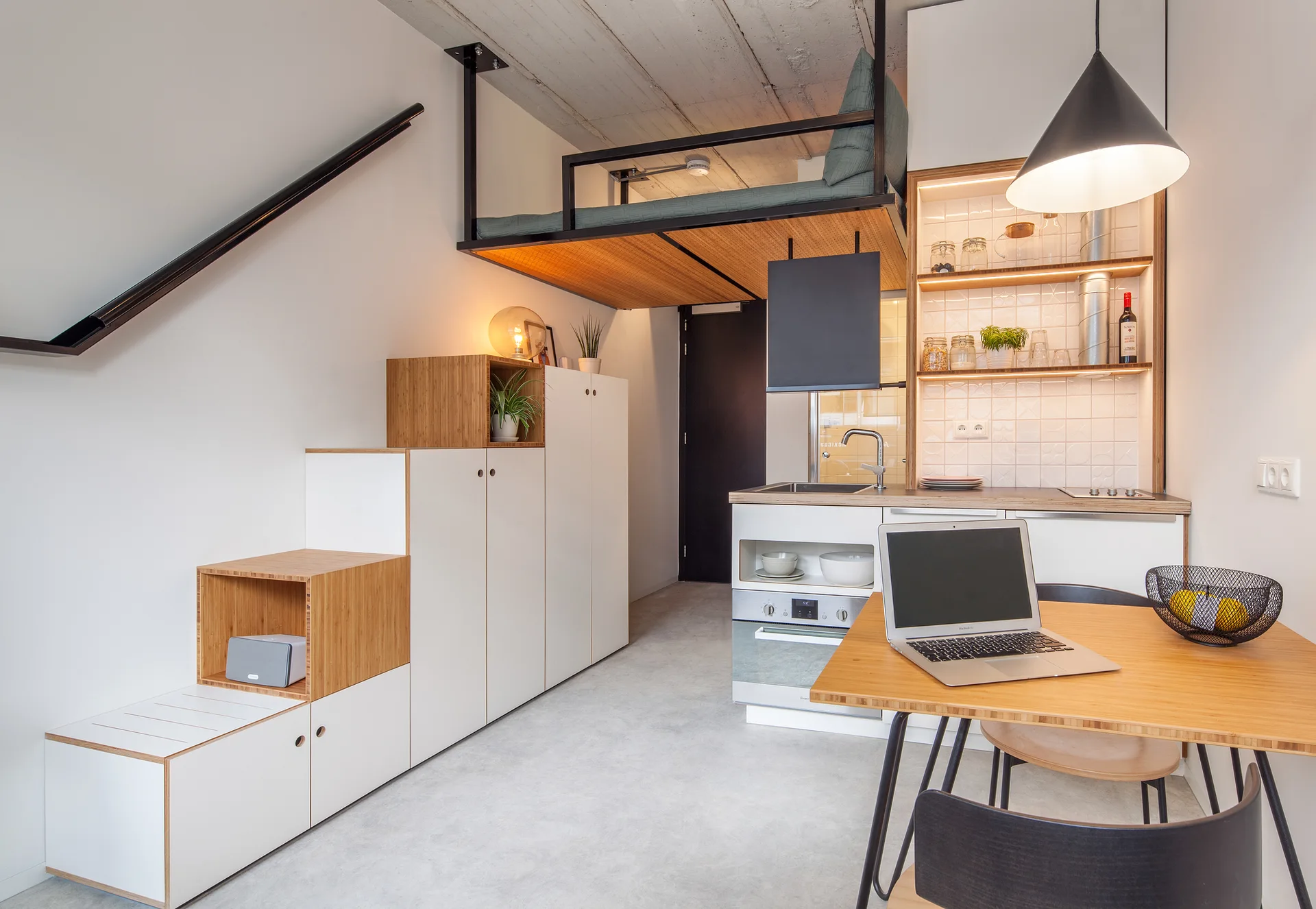A compact studio apartment features white kitchen cabinets, a wooden dining table, a metal-framed loft bed, and stepped storage cabinets.