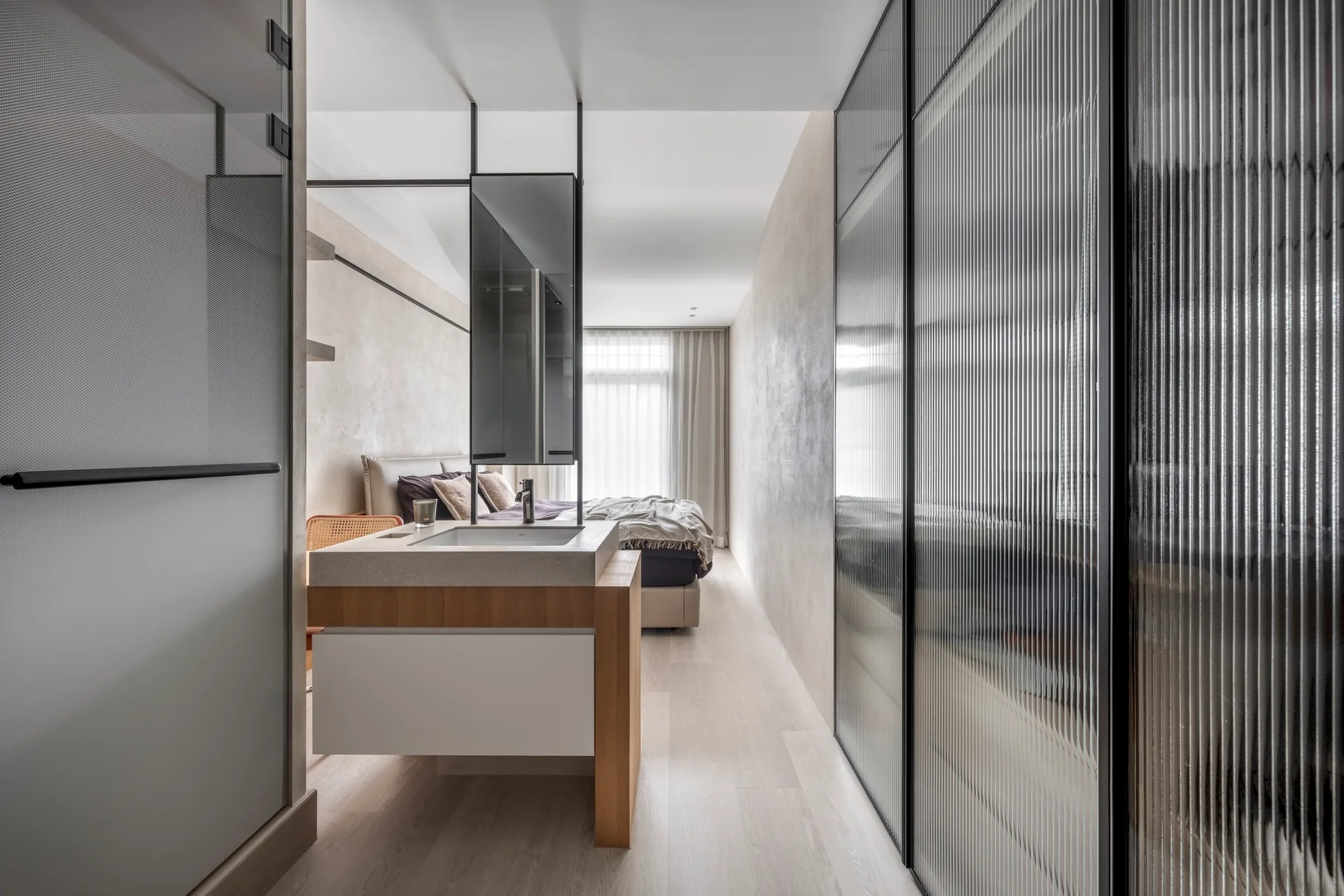 An open-concept bedroom features a freestanding light wood vanity with an integrated sink, plaster walls, and a wardrobe with fluted glass doors.