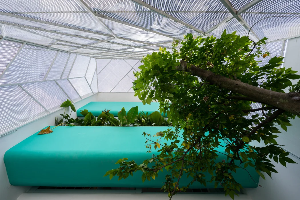 A modern courtyard features a large curved teal seating bench beneath a geometric perforated metal canopy with an integrated leafy tree.