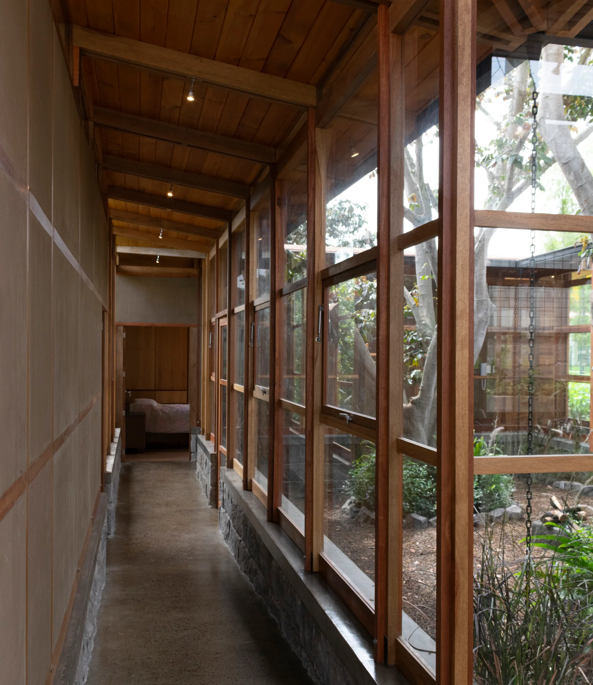 A concrete floor corridor features a wood plank ceiling and a continuous wood-framed window wall facing a landscaped courtyard garden.