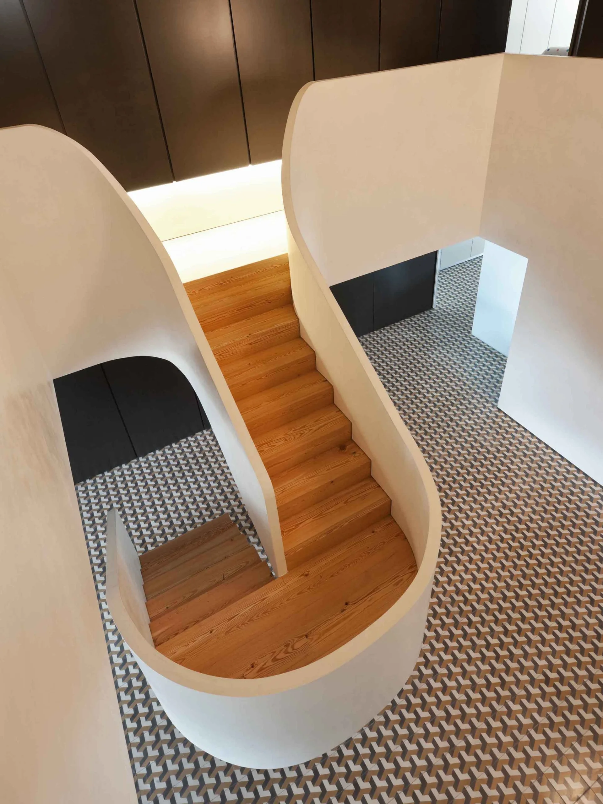 A top-down view of a curved white spiral staircase with light wood treads winding down to a geometric patterned tile floor.