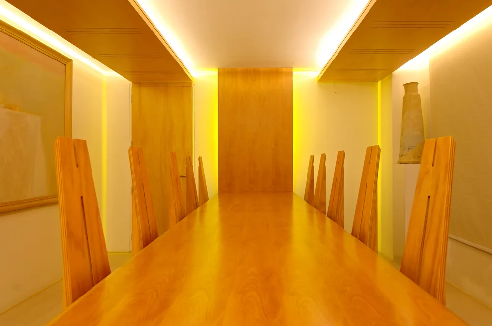 A long wooden dining table is flanked by tall, angular chairs in a minimalist room featuring warm cove lighting and backlit wood wall panels.