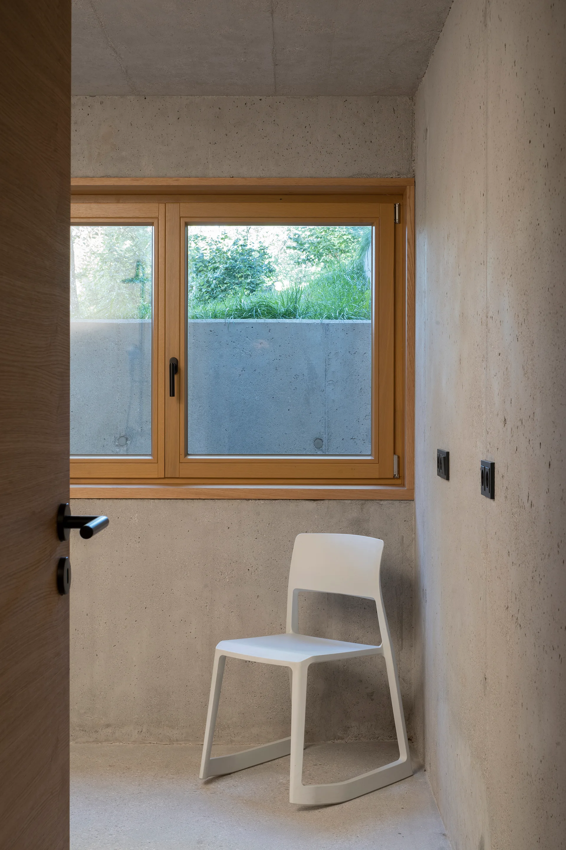 A white modern chair stands in a minimalist room with exposed concrete walls and a natural wood-framed window.