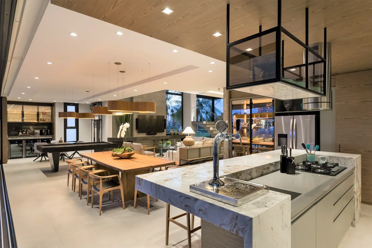 An open-concept kitchen and dining area features a veined marble island, a wooden dining table under pendant lights, and a wood-paneled ceiling.