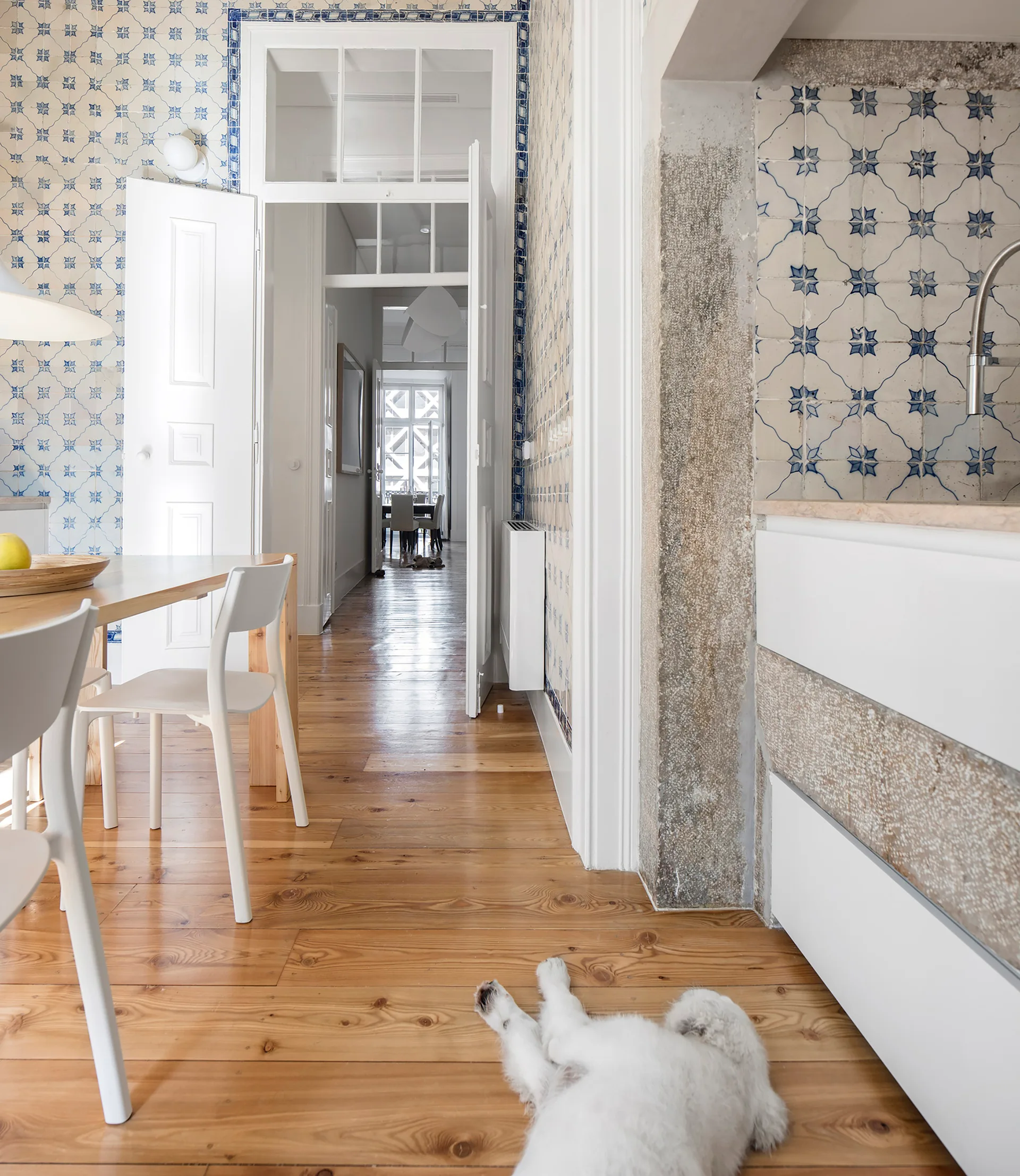 An interior kitchen and dining area featuring blue and white patterned wall tiles, light wood floors, and modern white cabinetry.