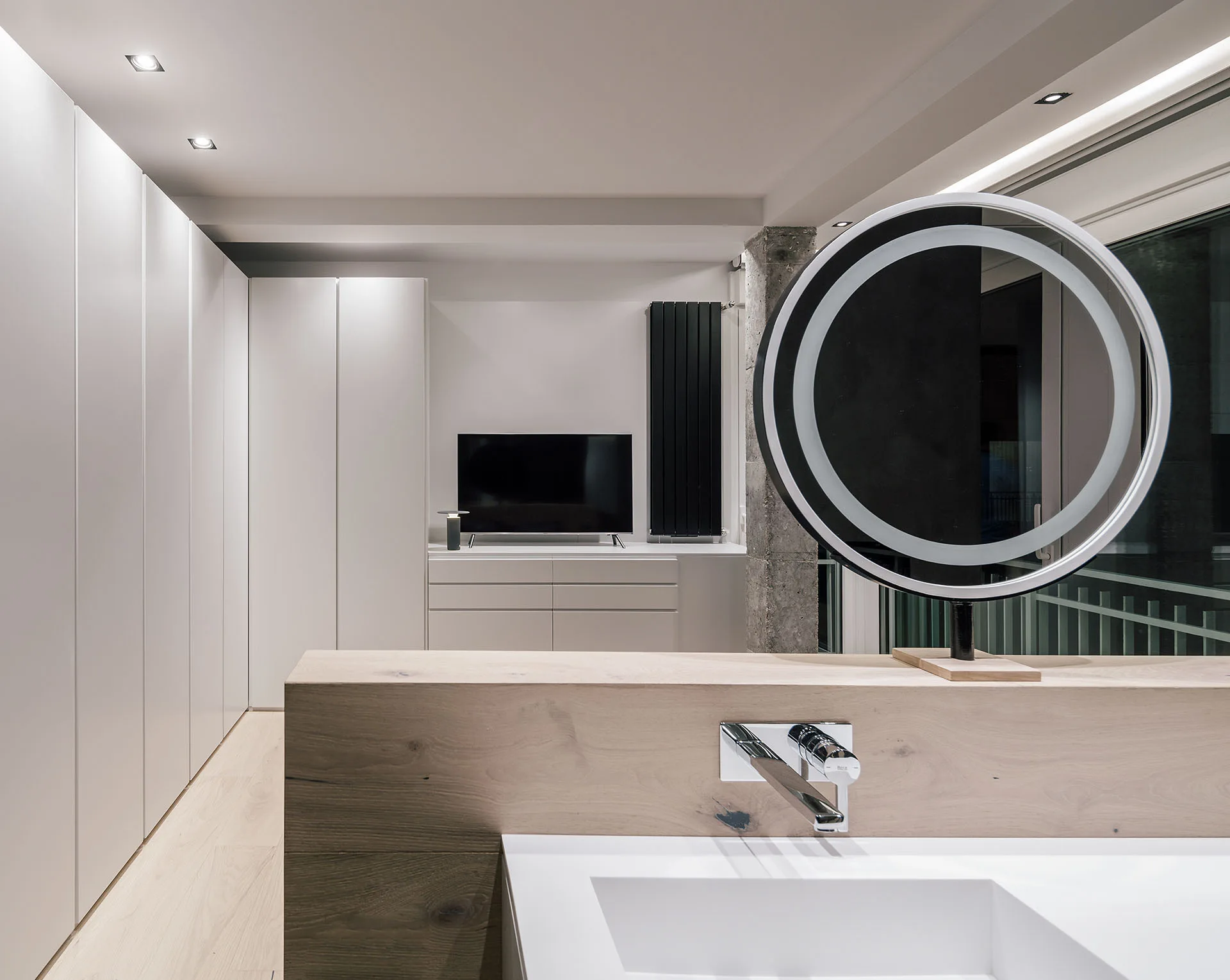 A light wood vanity partition with an integrated white sink, a chrome wall-mounted faucet, and a round illuminated LED mirror in a modern room.