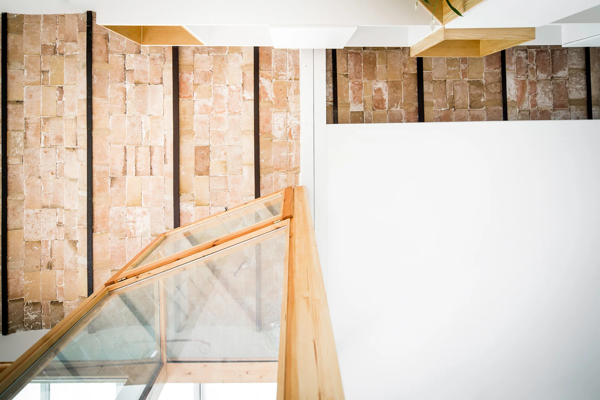 A high-angle view of a modern stairwell featuring an exposed brick ceiling with black metal beams and a light wood and glass staircase railing.