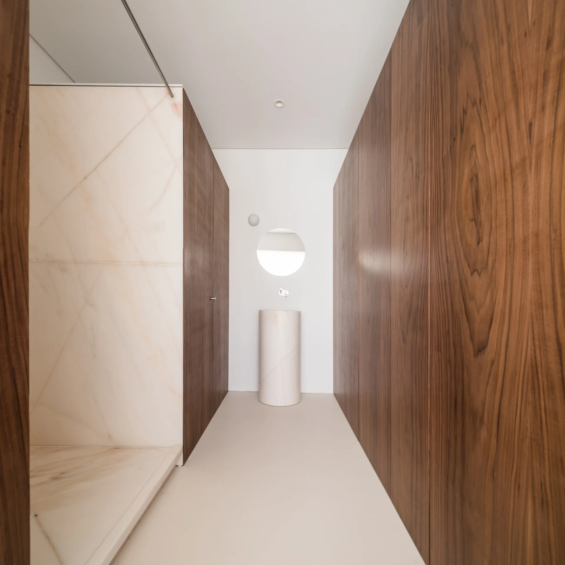 A minimalist bathroom corridor featuring full-height dark wood wall paneling, a marble walk-in shower, and a cylindrical marble pedestal sink.