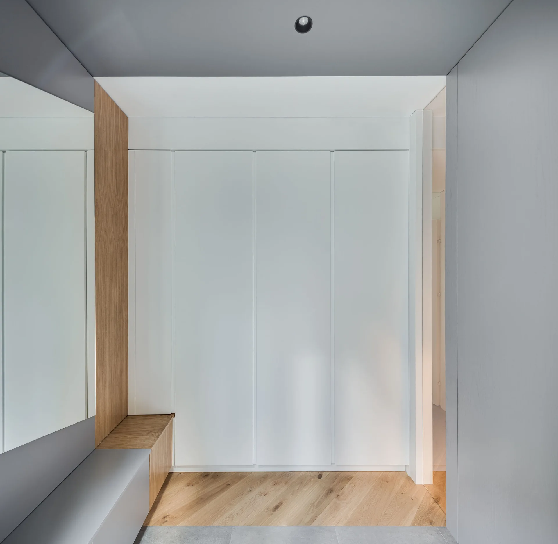 A minimalist entryway features seamless white built-in cabinets, a large wall mirror, a wood and grey bench, and diagonal wood flooring.