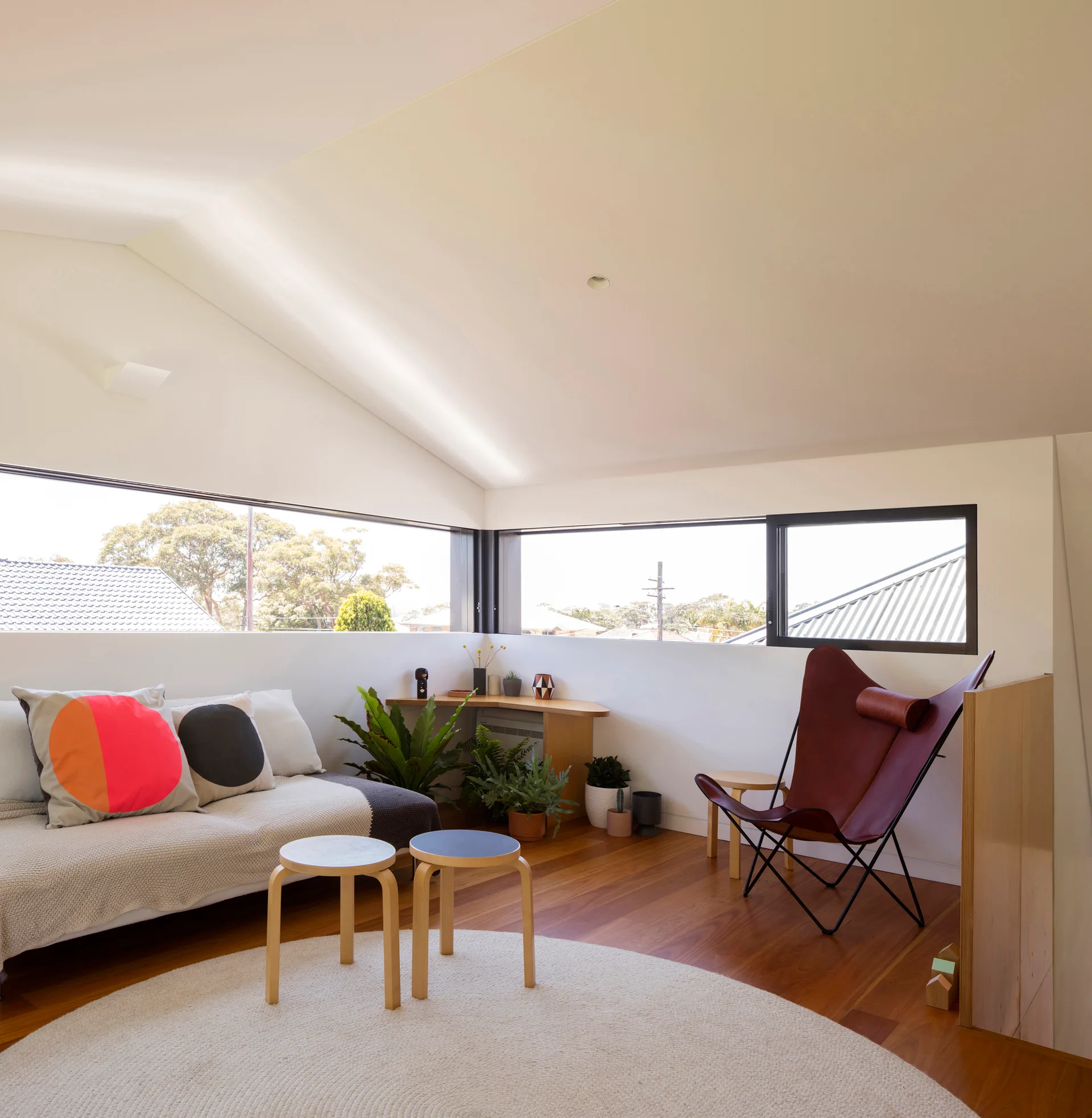 A loft living room features a low sofa with patterned pillows, a brown leather butterfly chair, wooden side tables, and horizontal clerestory windows.