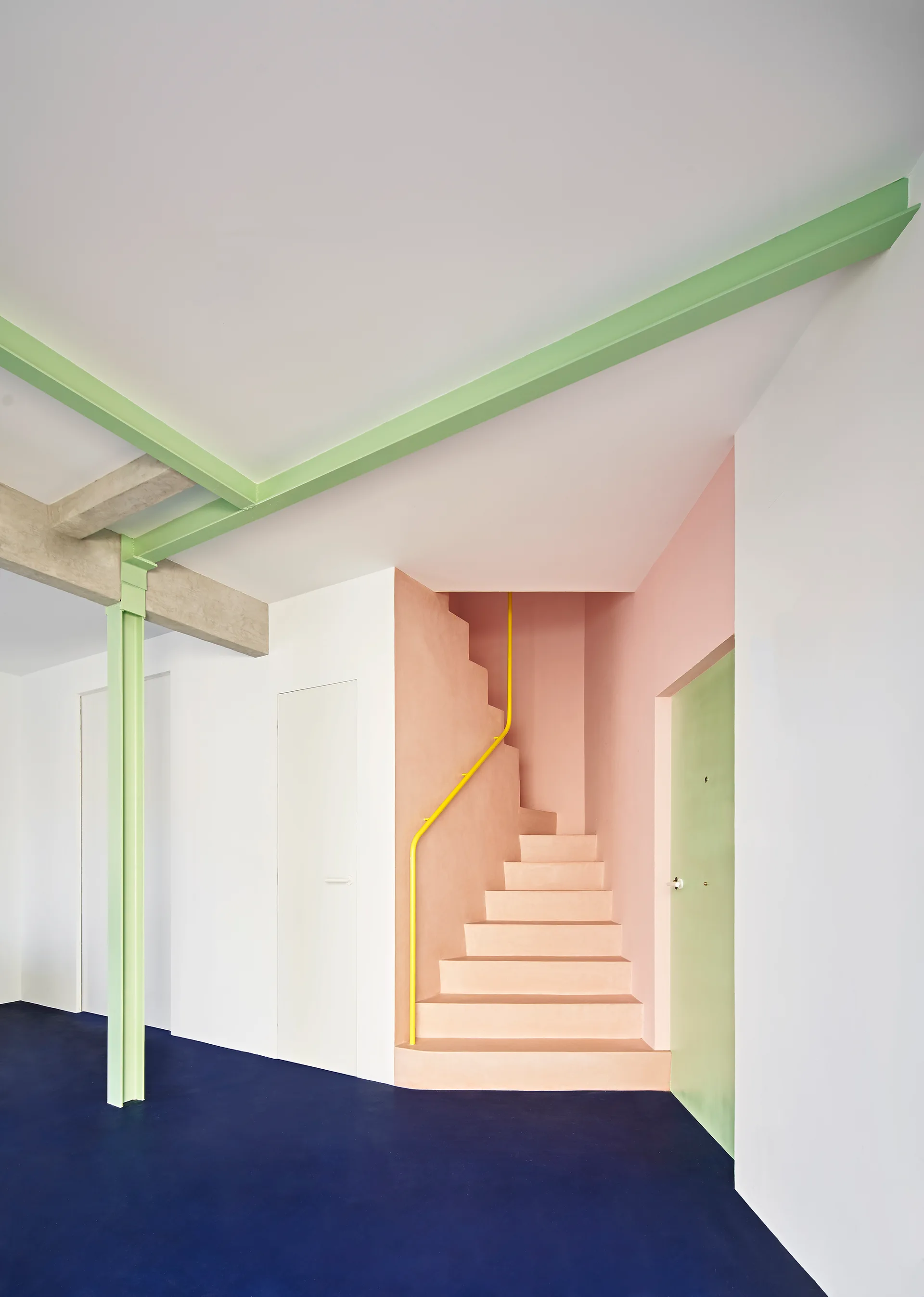 A pink minimalist staircase with a yellow handrail sits next to a green door, set against a deep blue floor and light green structural beams.